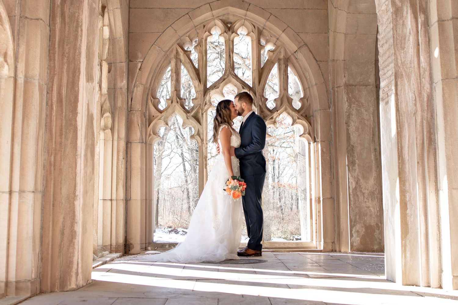 A bride and groom embrace in front of a stunning Gothic arched window in an elegant stone cathedral setting.