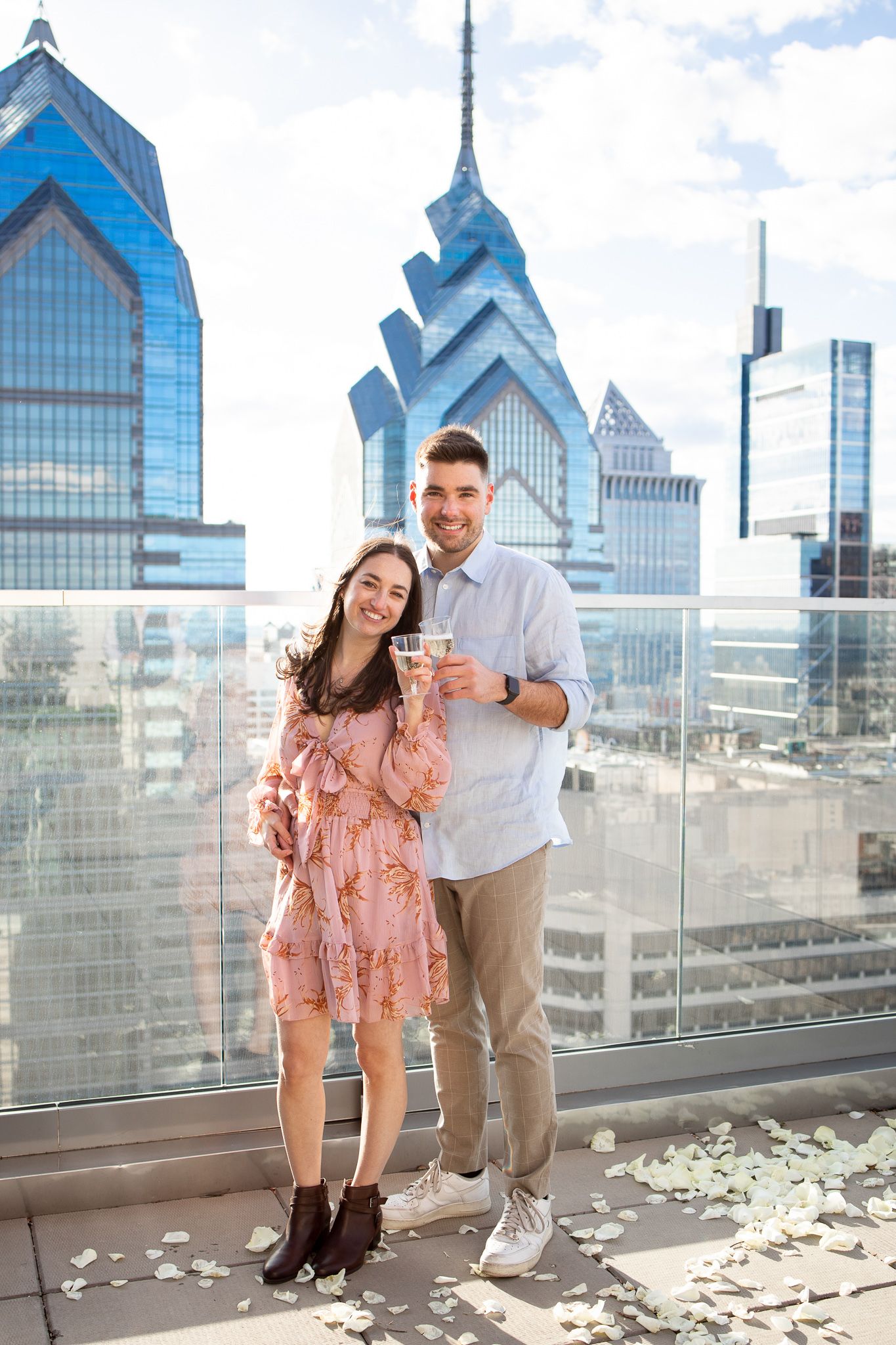 A couple shares a romantic moment on a rooftop terrace overlooking the Philadelphia skyline with rose petals scattered around.