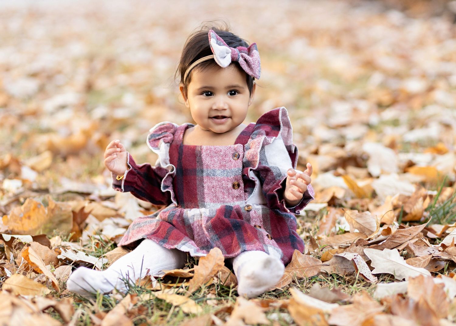 Small child wearing a pink and burgundy dress sits among fallen autumn leaves on the ground.