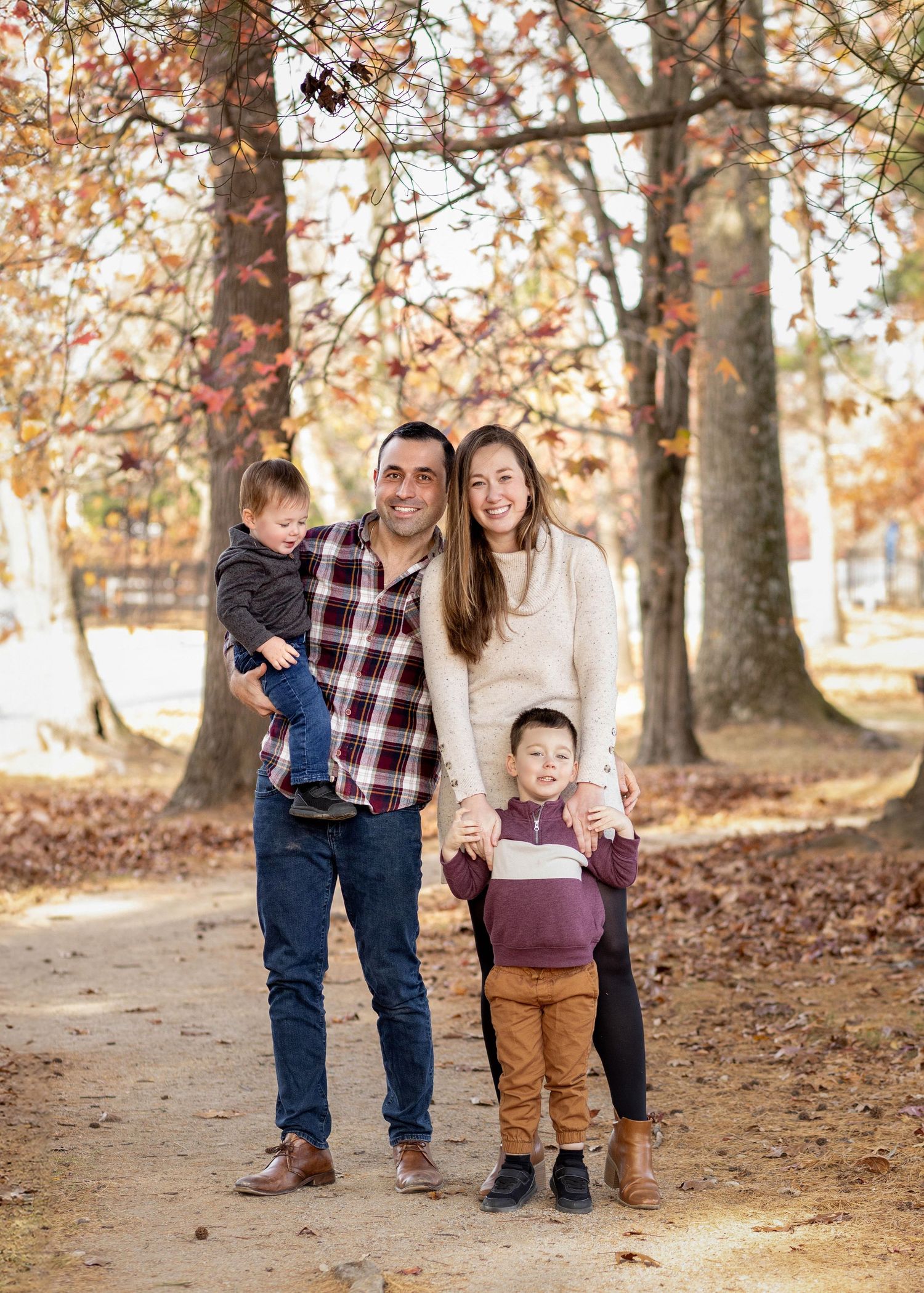 Family poses together on a leaf-covered path in the woods during autumn with fall foliage in the background.