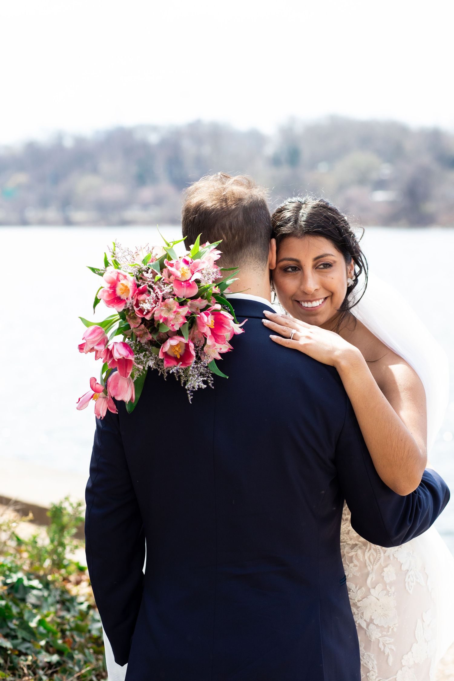 A couple embraces on their wedding day while holding a vibrant pink floral bouquet near a waterfront.