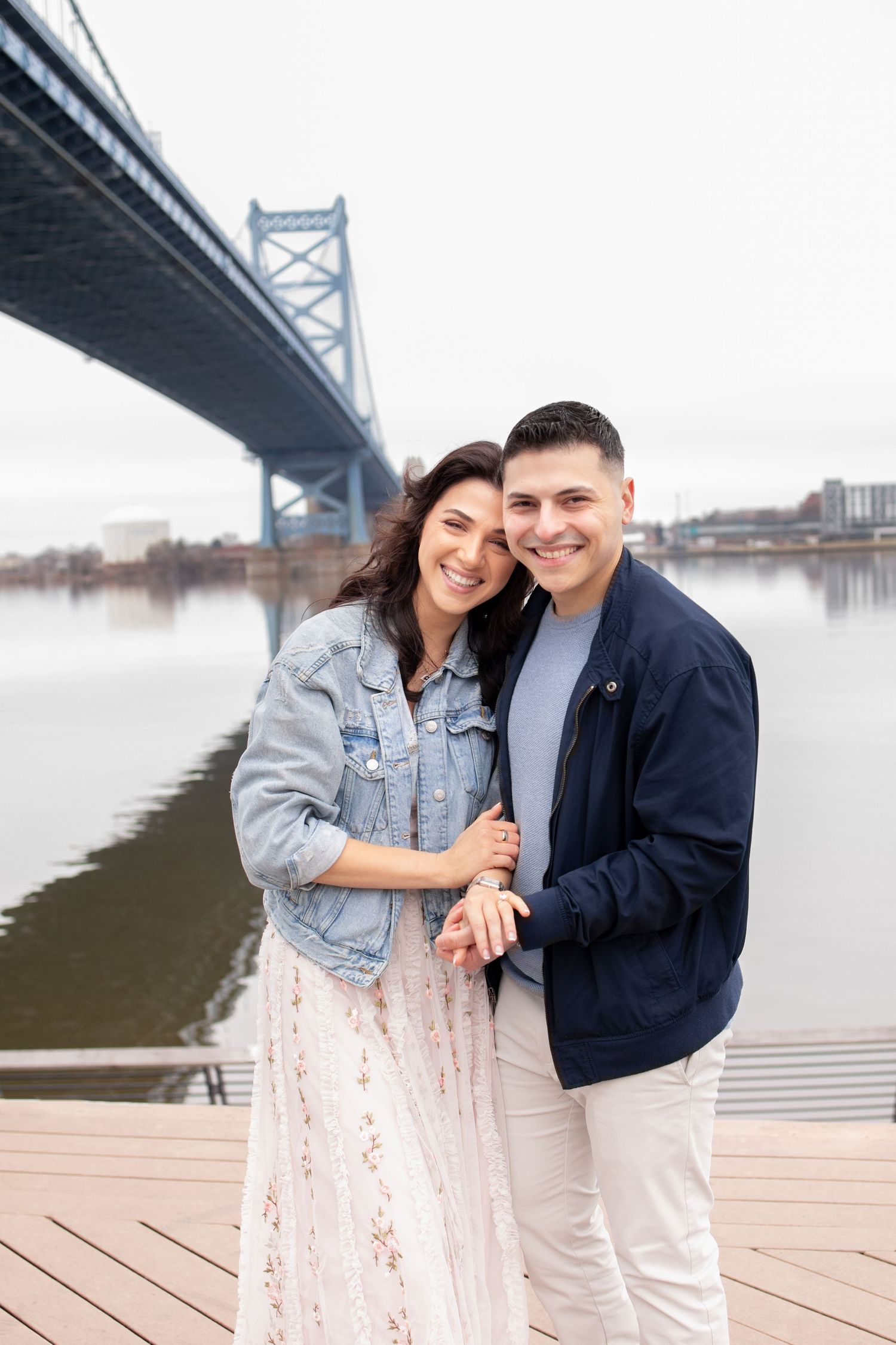A joyful couple embraces while standing on a wooden deck near the Benjamin Franklin Bridge in Philadelphia.