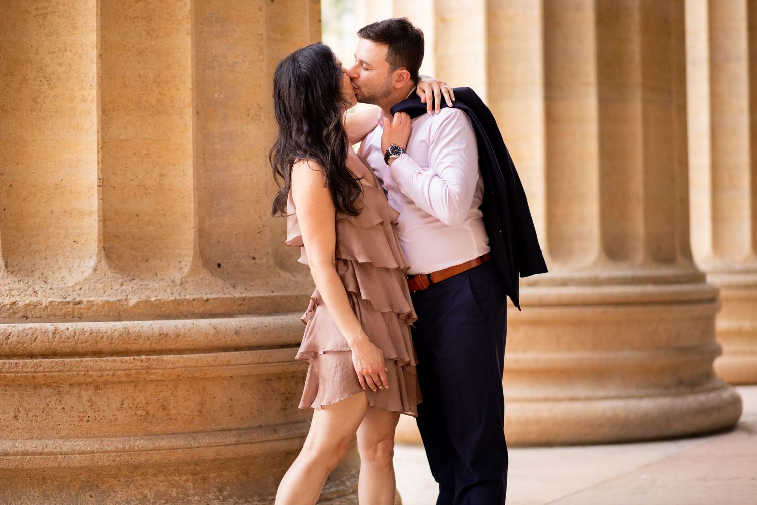 A romantic couple embraces and kisses against elegant stone pillars, with the woman wearing a blush fringe dress.