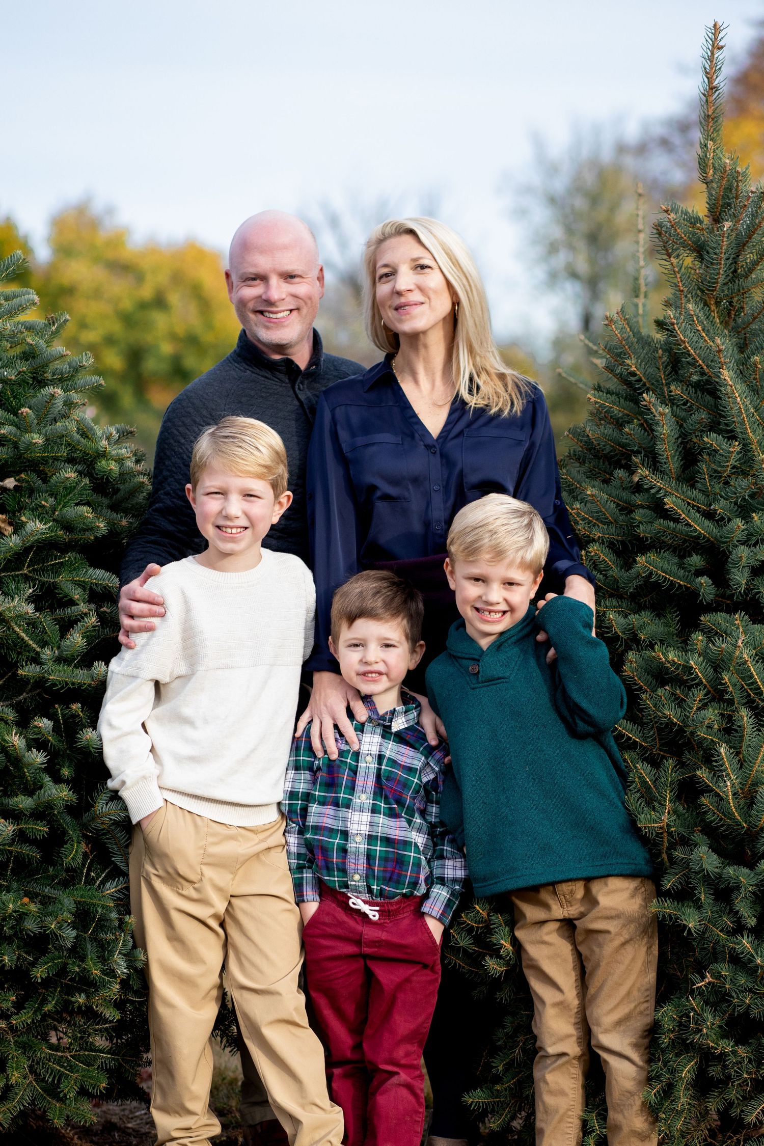A family poses together among evergreen Christmas trees during a festive outdoor holiday photo session.