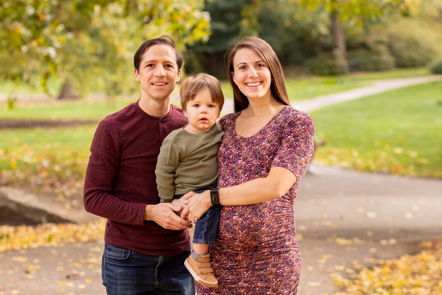 Happy family of three posing outdoors in a park during autumn, with colorful fall foliage in the background.