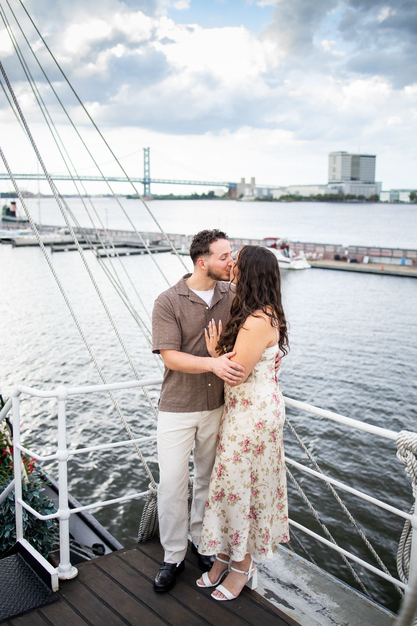 A couple shares a romantic moment on a dock overlooking a scenic harbor with boats and cloudy skies in the background.