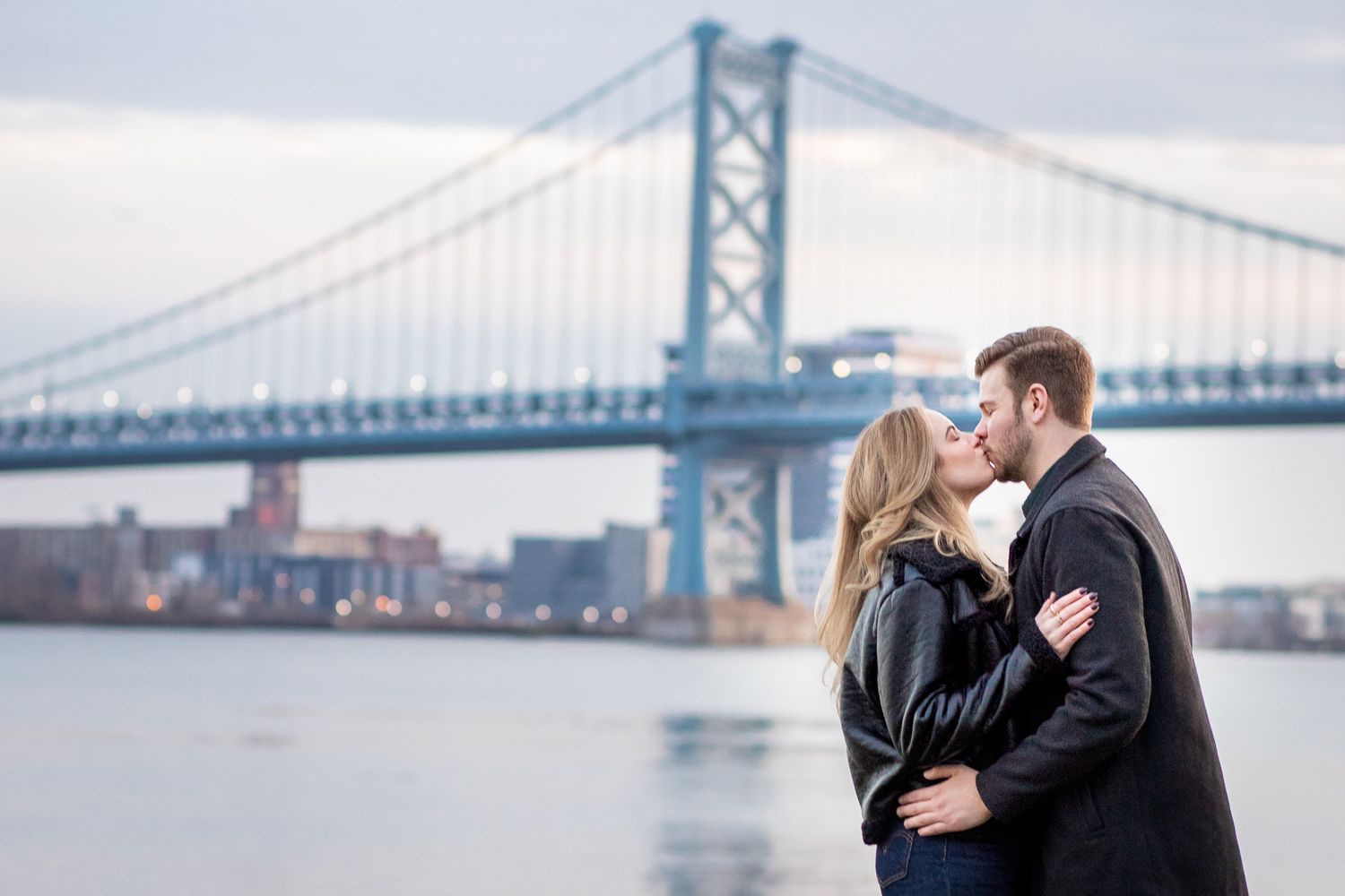 A couple shares romantic moments by the Benjamin Franklin Bridge in Philadelphia at dusk.
