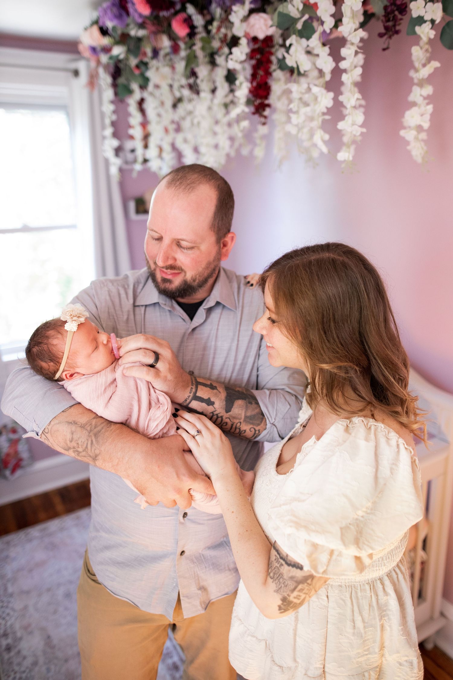 In a pink floral nursery, parents hold and gaze lovingly at their newborn baby while standing together under hanging flowers.