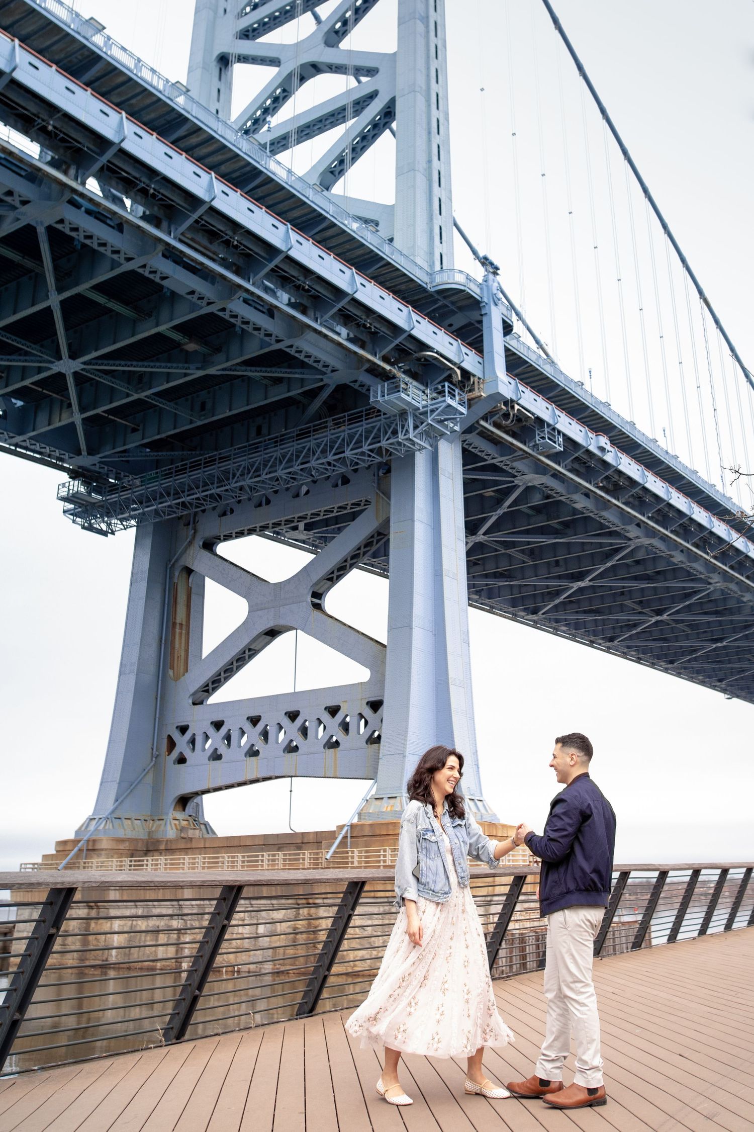 A romantic couple enjoys a moment together on a wooden pier beneath the massive steel structure of a bridge.