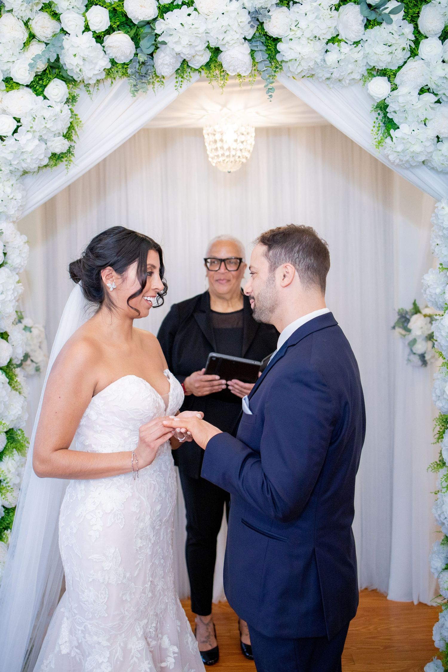 Wedding ceremony takes place under white floral arch with crystal chandelier and draped fabric decorations.