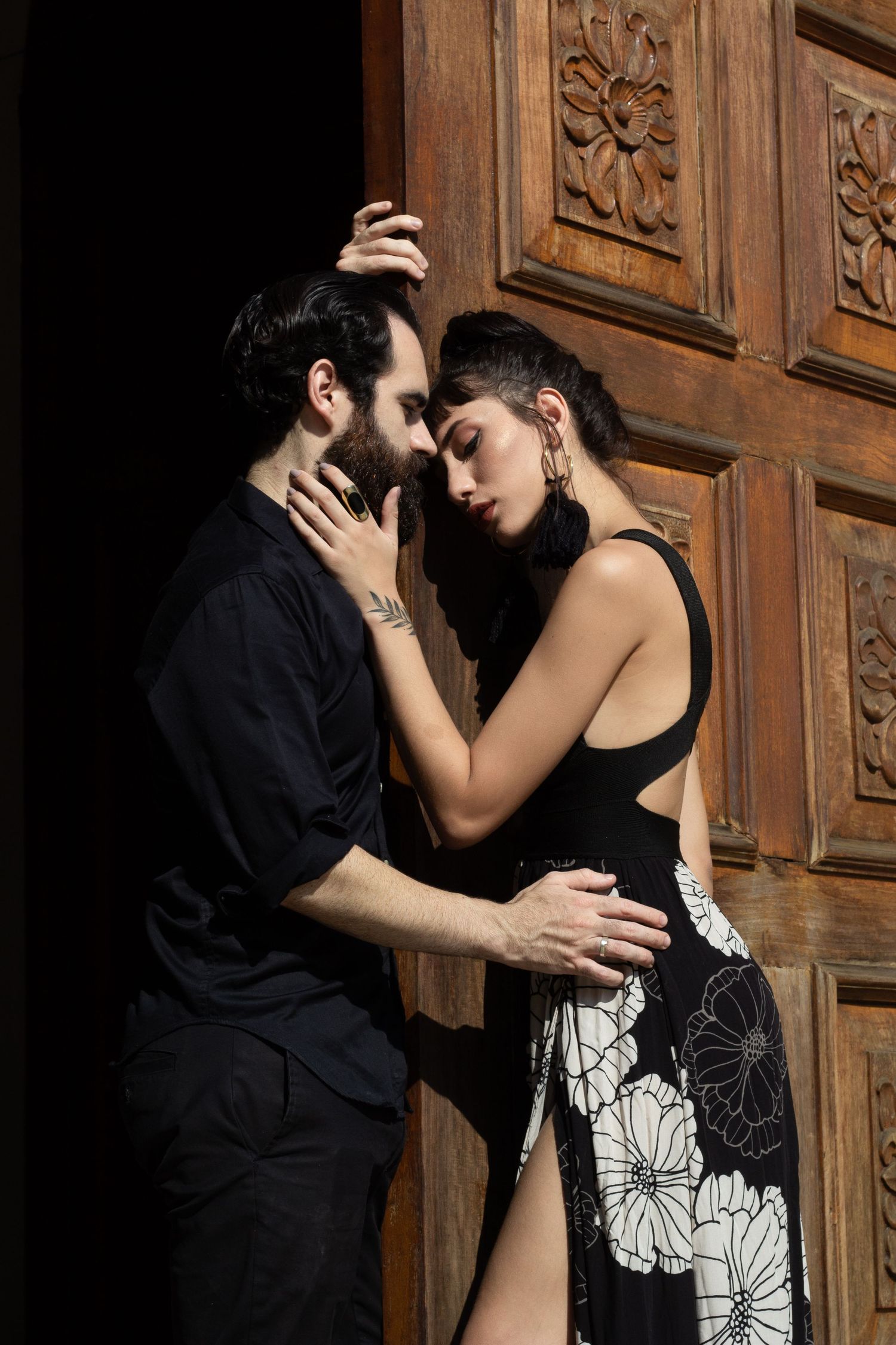Romantic couple embraces against ornate wooden door in dramatic lighting.