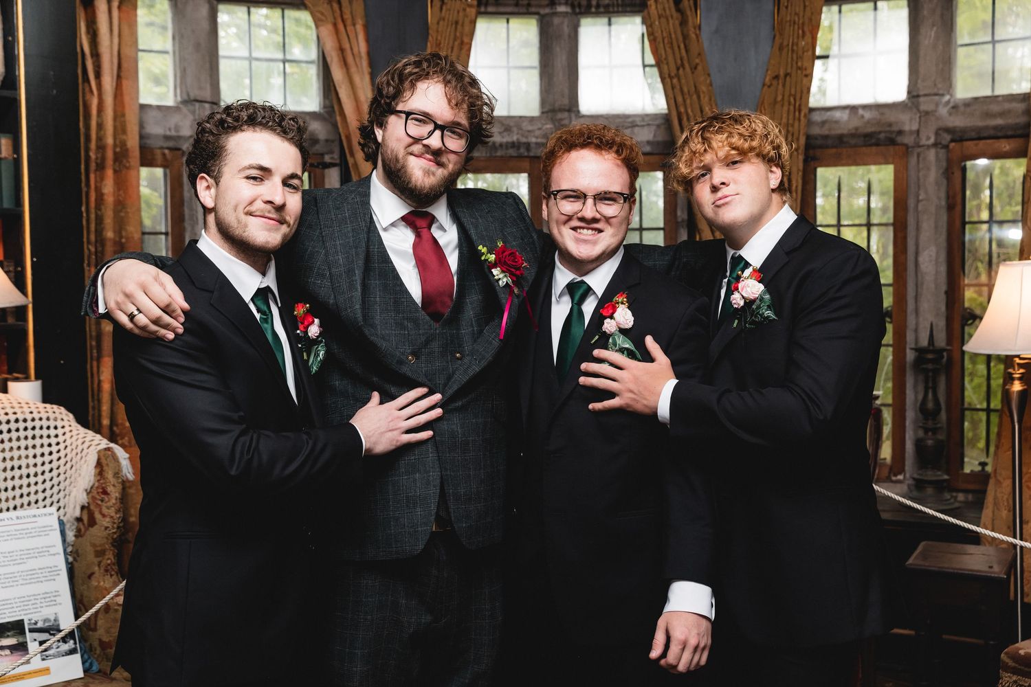 Four groomsmen in black tuxedos and red ties pose together while smiling at a wedding reception.