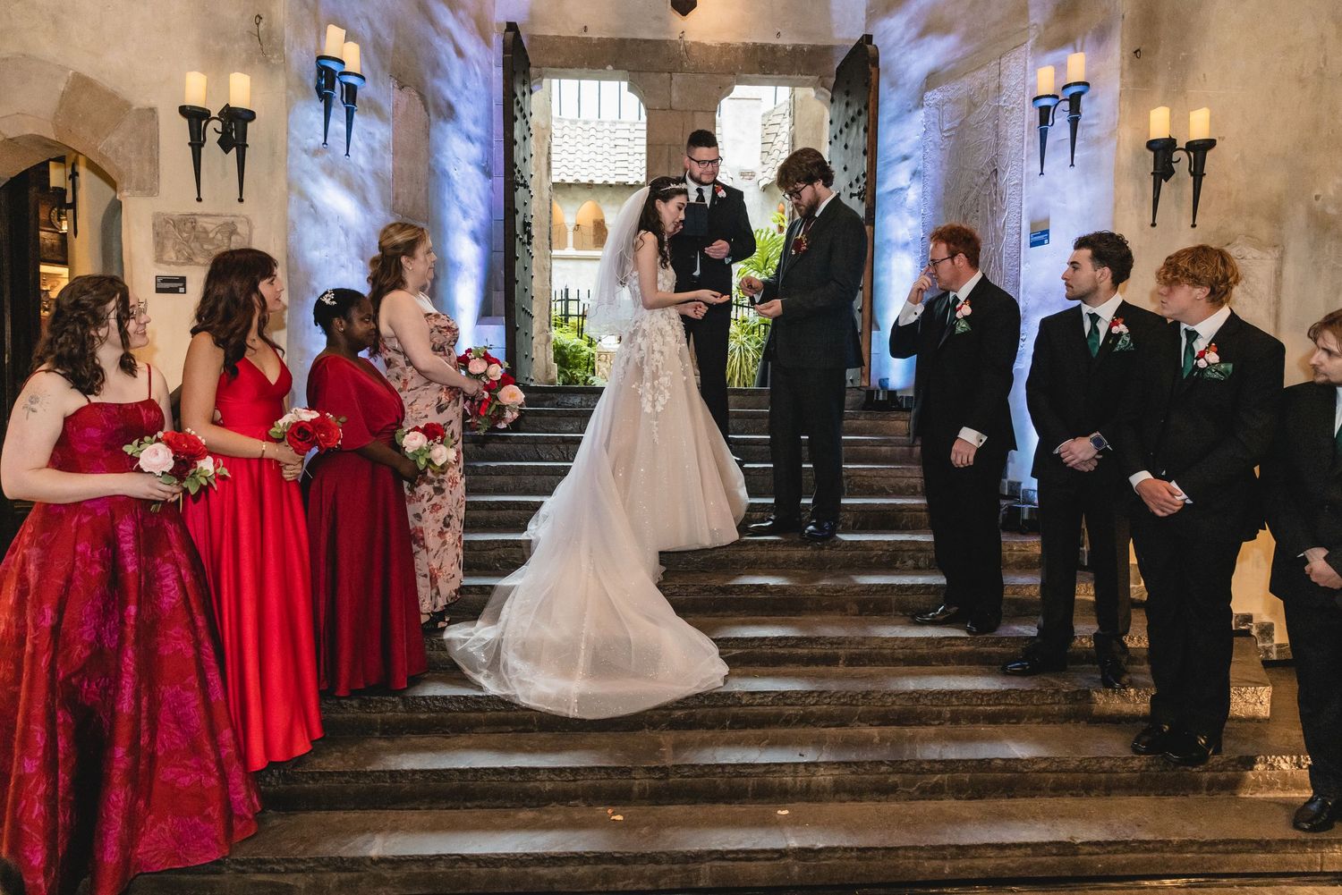 A wedding ceremony in a grand stone chapel with bridesmaids in red dresses and groomsmen in black suits lined up on each side.