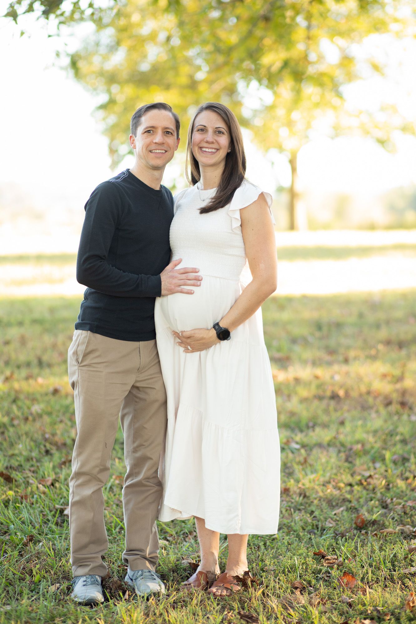 A happy couple poses outdoors, the woman in a white dress cradling her pregnant belly in a sunny park.