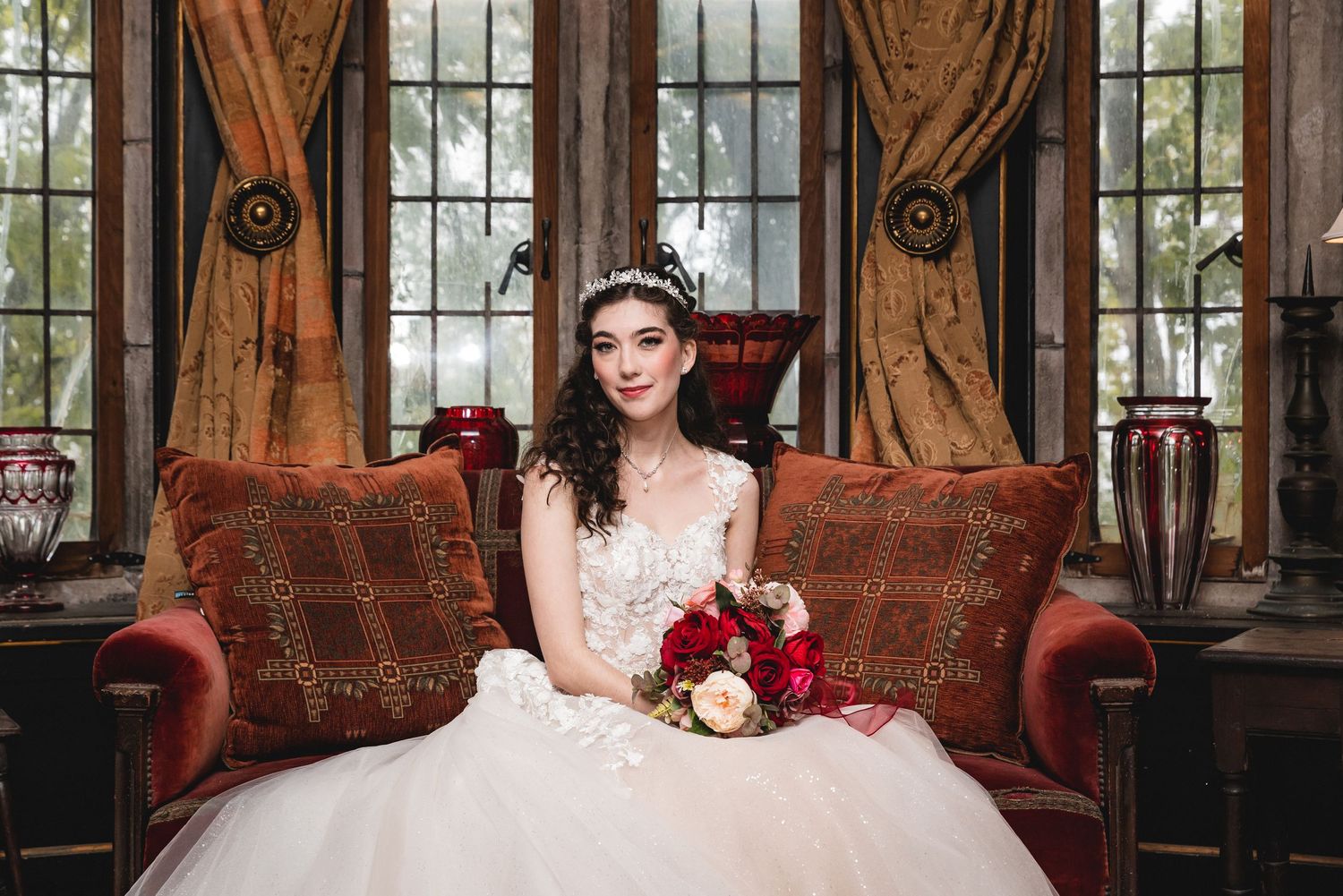 A bride in a white ballgown sits on an antique red couch holding a deep red bouquet in an elegant conservatory setting.