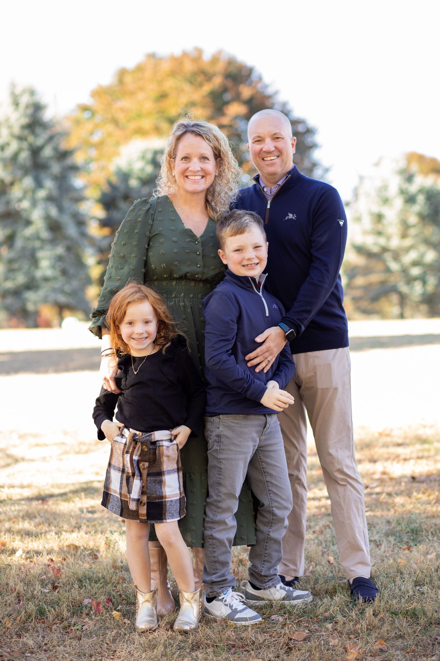 A family poses together outdoors on an autumn day with fall foliage and evergreen trees in the background.