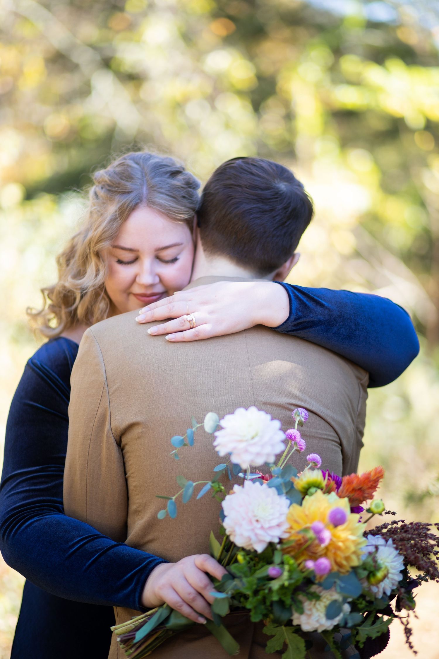 A couple embraces outdoors while holding a vibrant bouquet of flowers with pink and yellow blooms in autumn light.