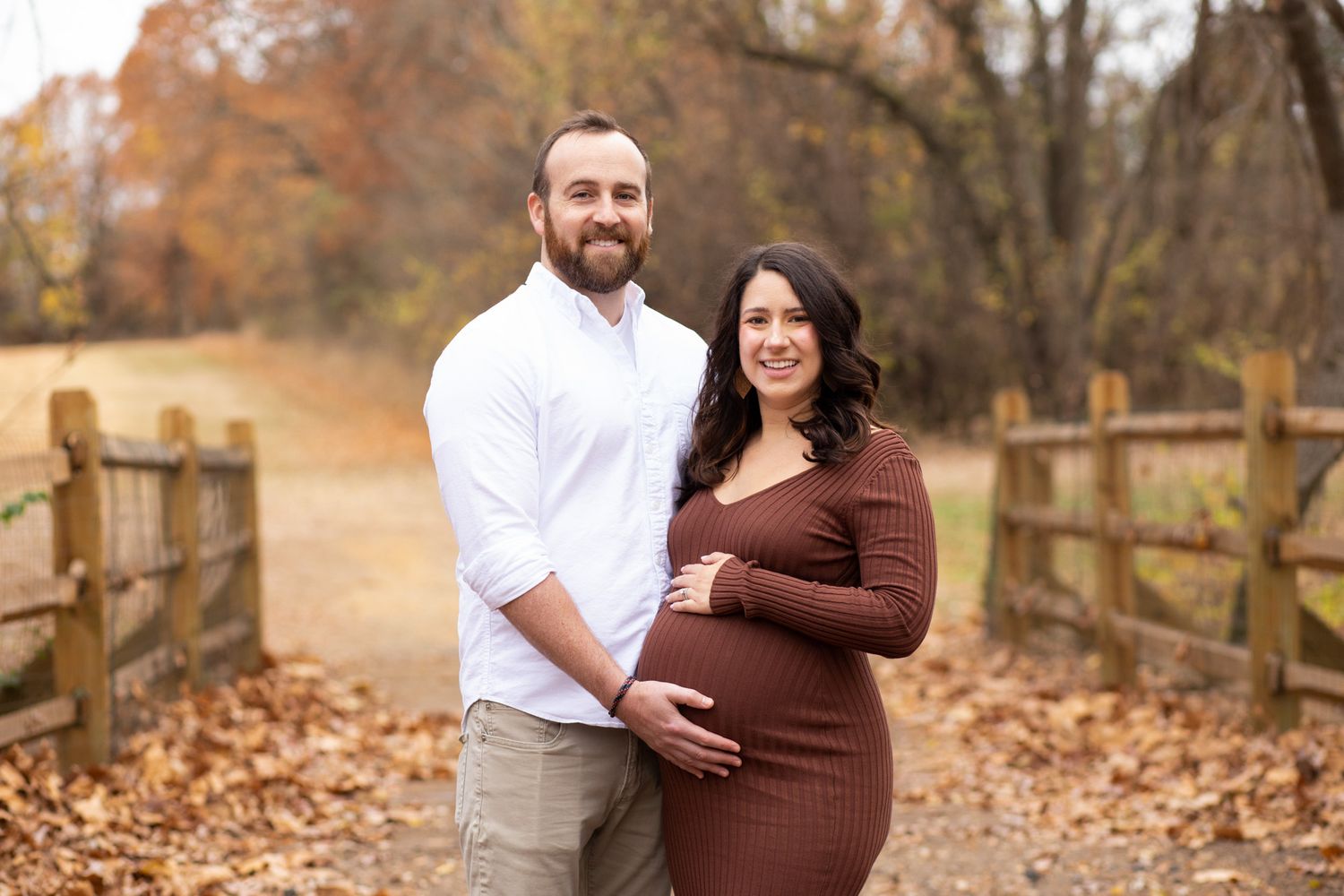 Couple poses outdoors in fall foliage, woman in brown maternity dress cradling baby bump, man in white shirt beside her.