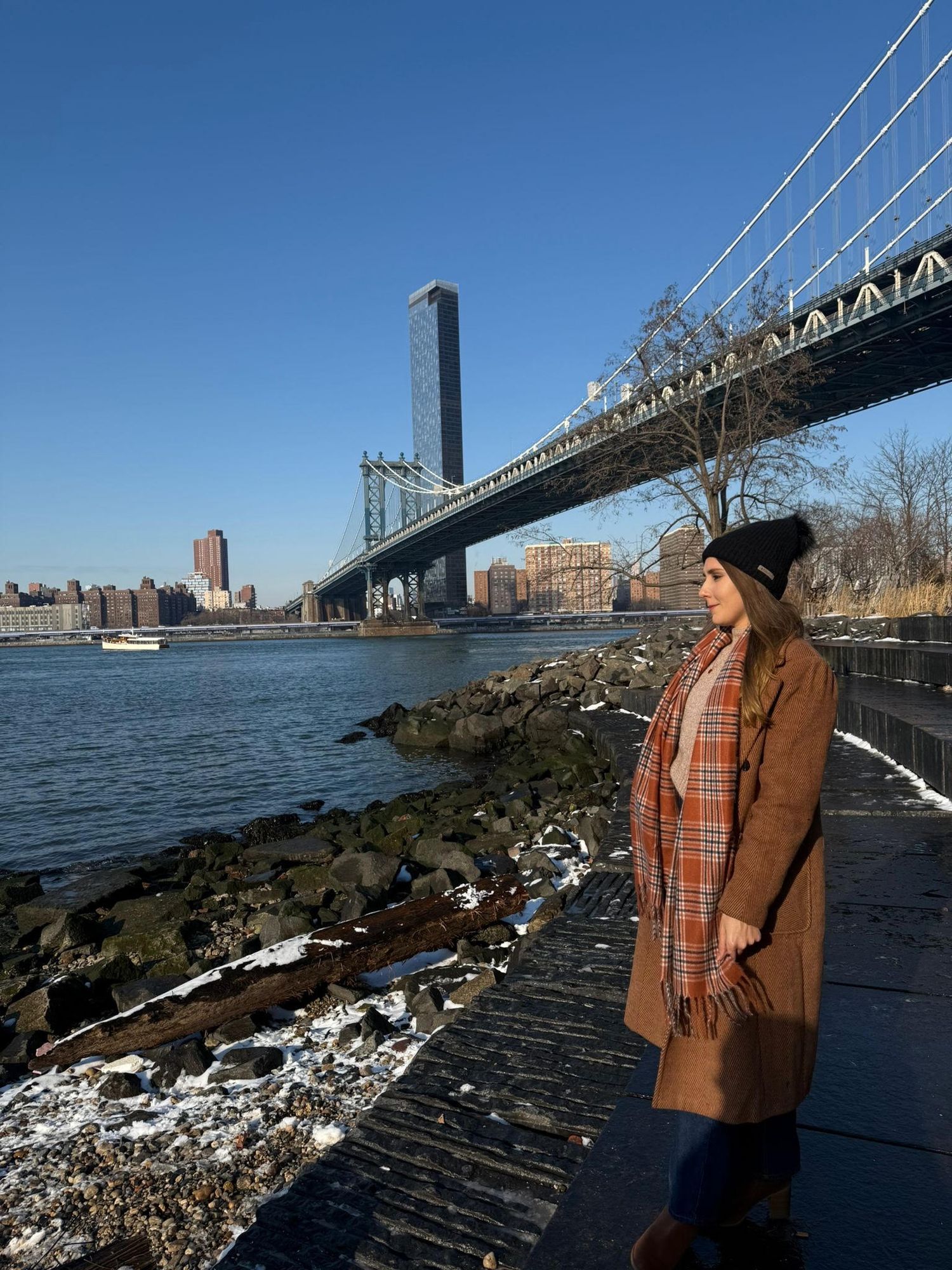 Someone in a tan coat and hat stands by the rocky shoreline under the Manhattan Bridge on a clear winter day in New York City.