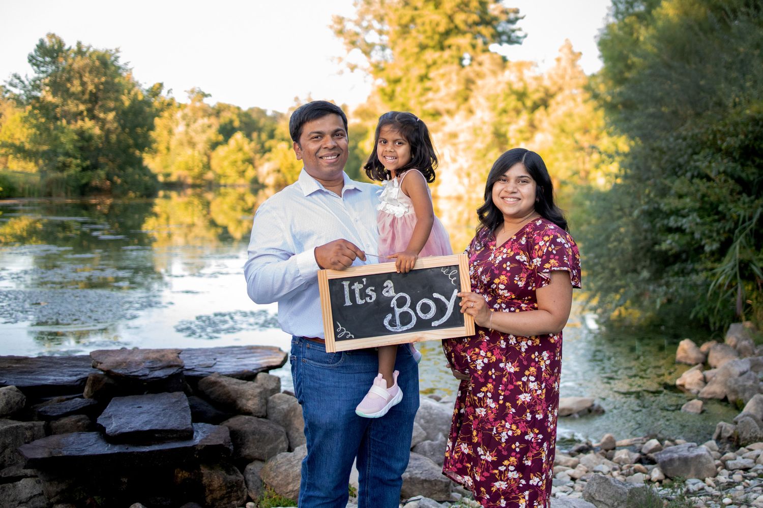 Family gender reveal photo with couple and toddler holding Its a Boy chalkboard sign outdoors.