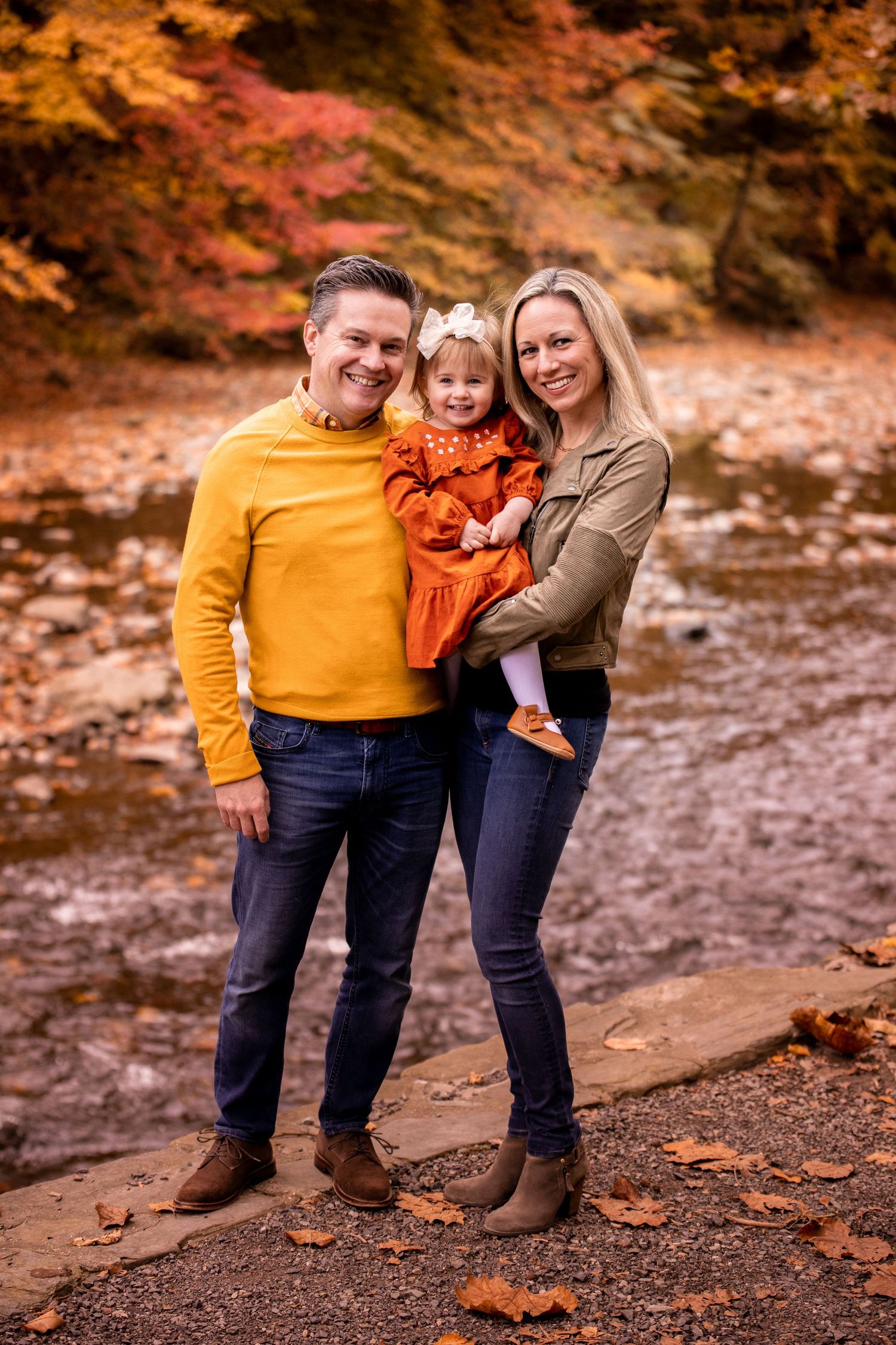 Three people in coordinating fall outfits pose together on a trail surrounded by autumn foliage with orange and red trees.
