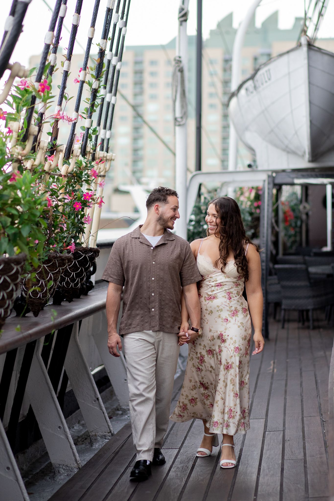 A couple walks hand in hand on a wooden deck with pink flowers and a boat in the background.