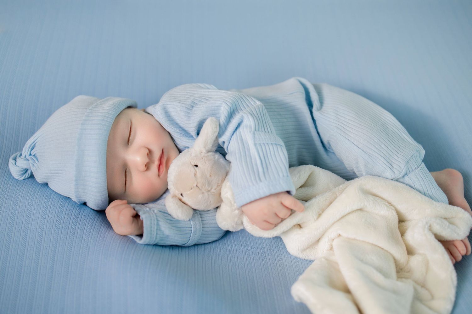 A sleeping baby in light blue clothing lies peacefully on a white blanket with a stuffed animal nearby.