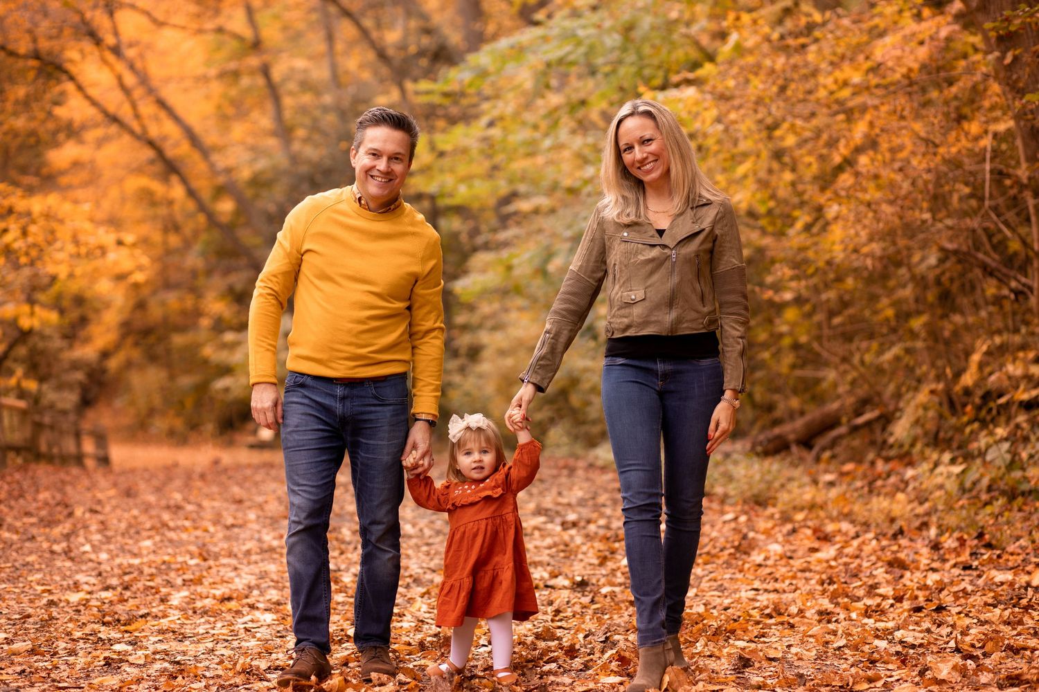 A family walks together while holding hands among fallen autumn leaves in a park surrounded by orange foliage.