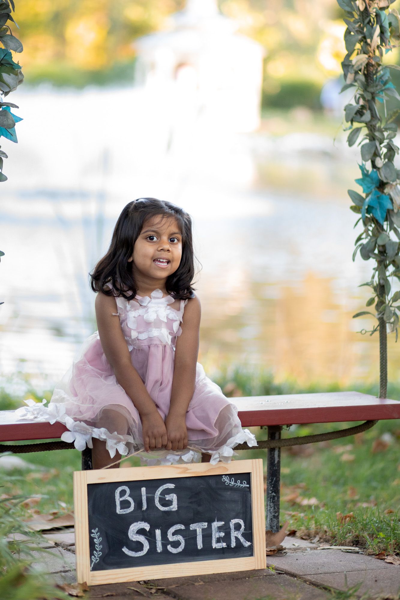 Young girl in pink dress sits on bench with Big Sister chalkboard sign, outdoor park setting with pond backdrop.