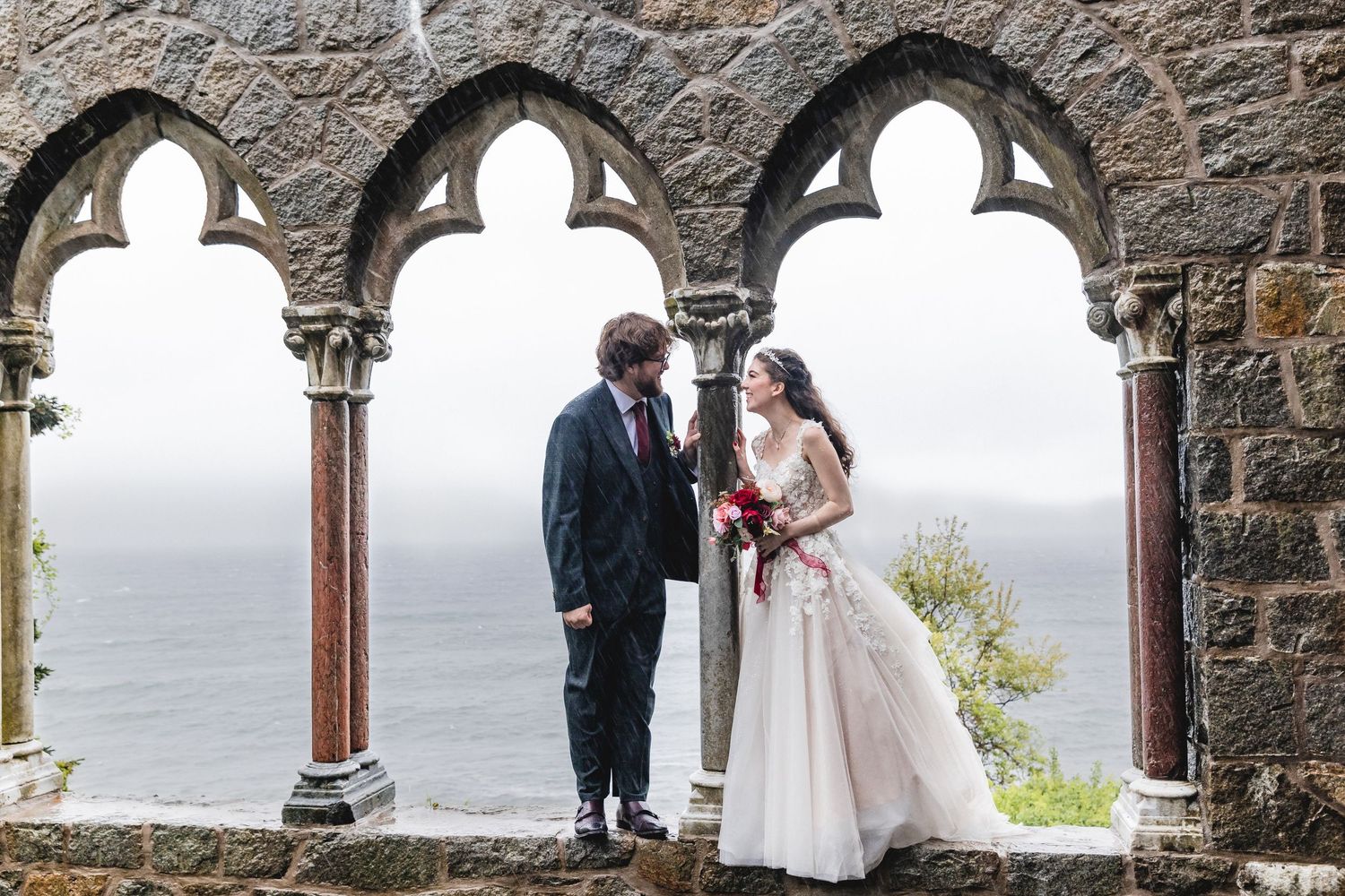 A couple shares a romantic moment under stone arches overlooking a misty coastline.