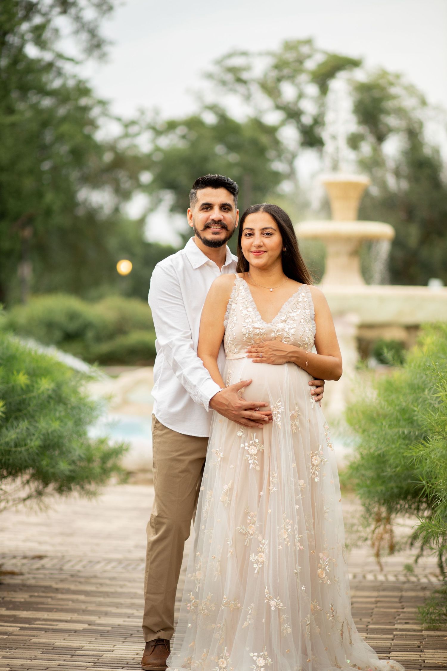 A couple stands outdoors near a decorative fountain as sunset light filters through trees in the background.