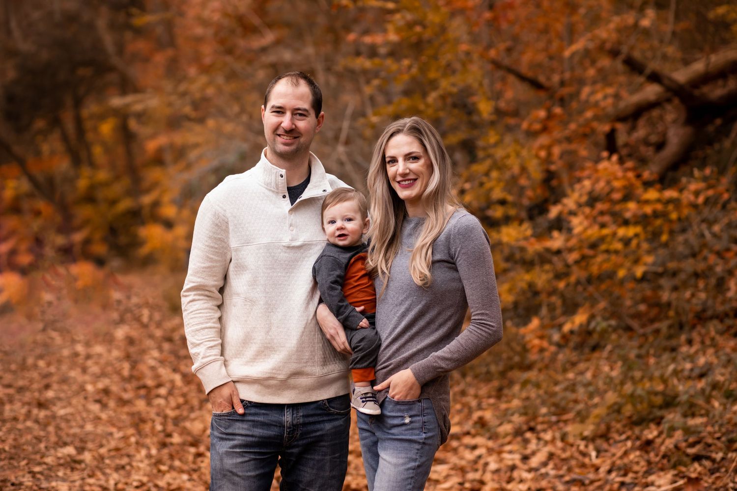 Family of three poses outdoors in casual clothing surrounded by vibrant autumn foliage and fallen leaves.