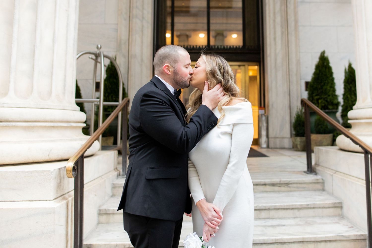 Couple shares intimate kiss on stone steps outside elegant building entrance with white pillars.