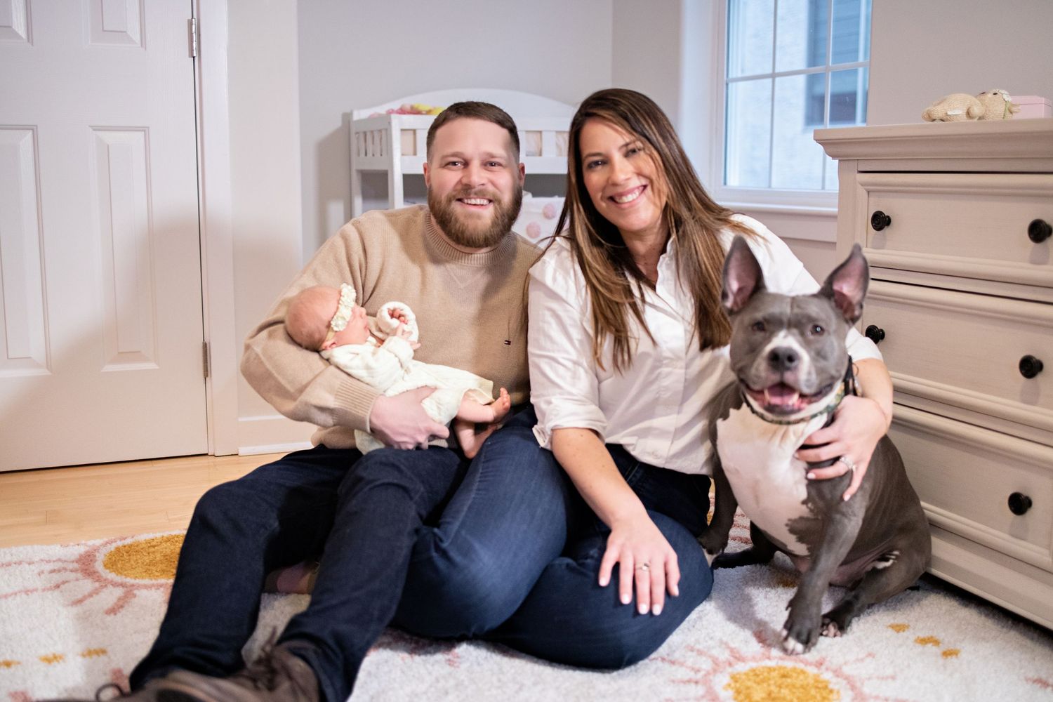 A couple sits on their floor at home with their white bunny and gray pitbull, surrounded by white furniture.