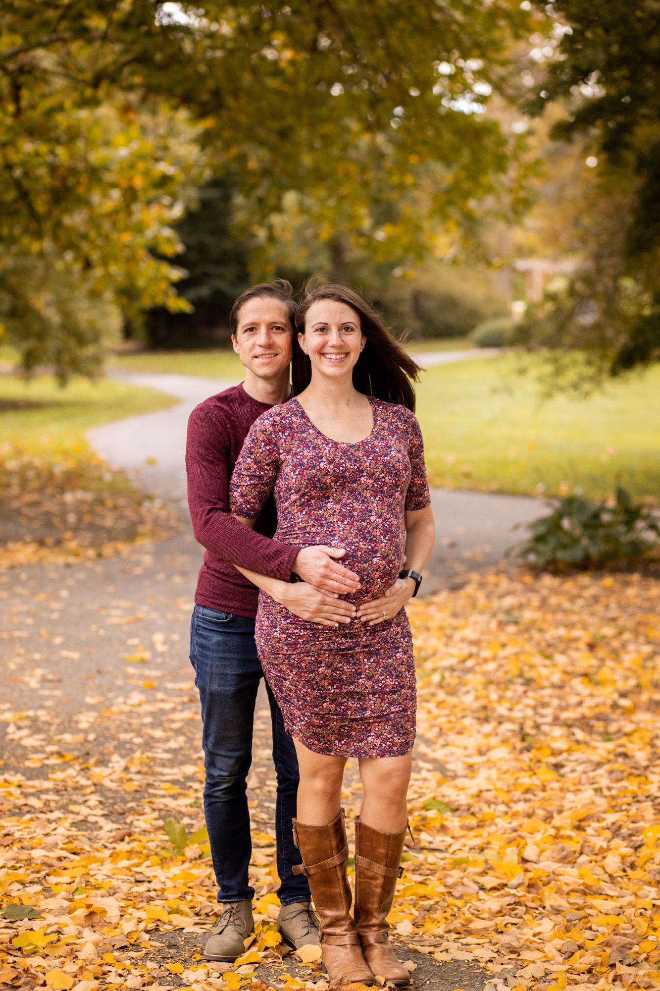 Couple posing in autumn park, man embracing pregnant woman in floral dress and boots, surrounded by golden fallen leaves.