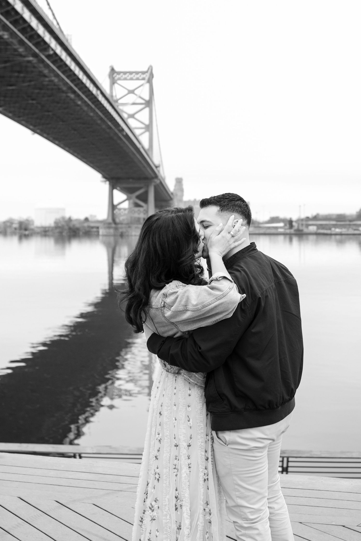 A romantic couple embraces and kisses by the Benjamin Franklin Bridge in Philadelphia with foggy views of the river and pier.