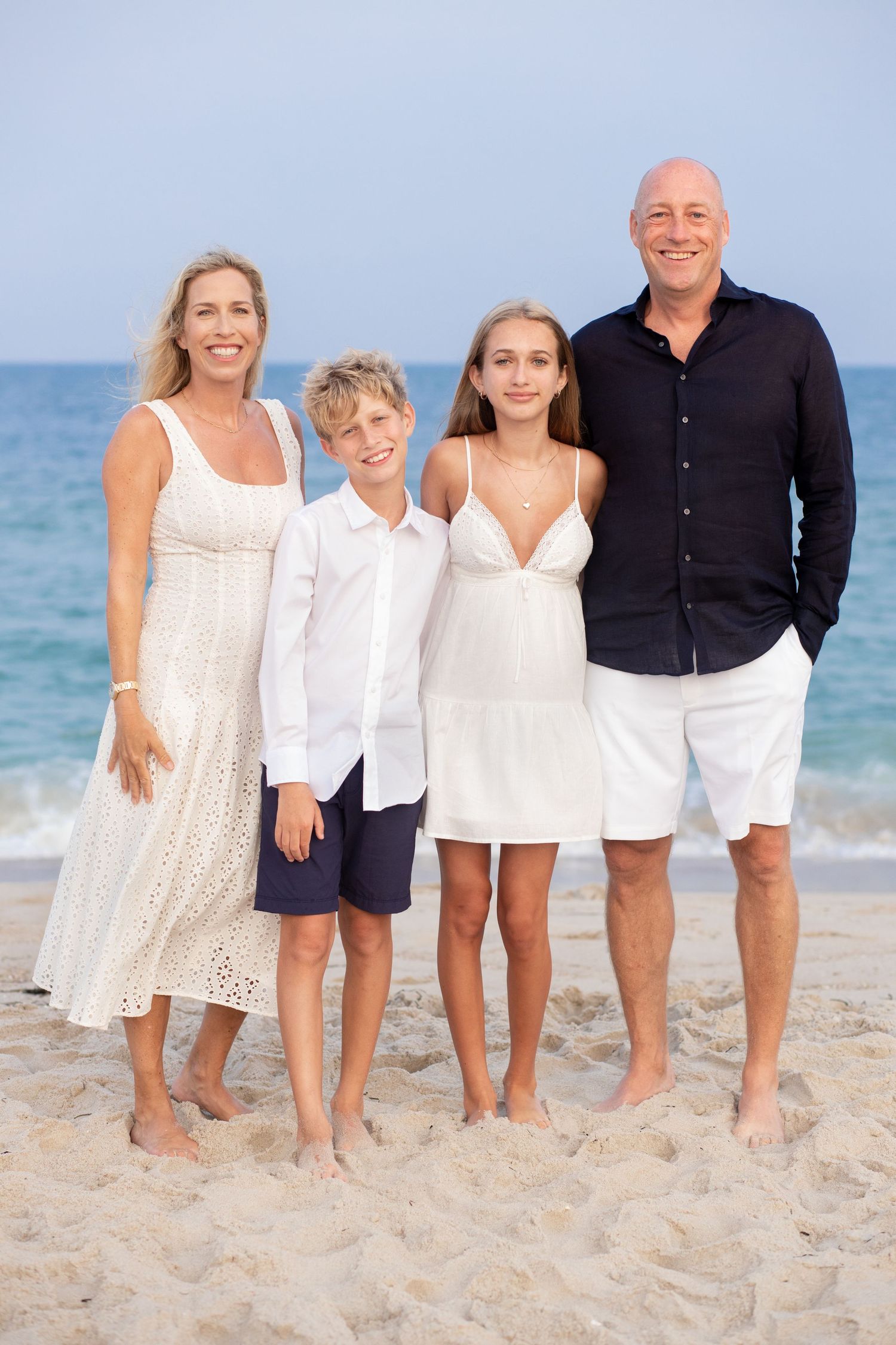 Four people dressed in white and navy casual attire pose together on a sunny beach with blue ocean in background.