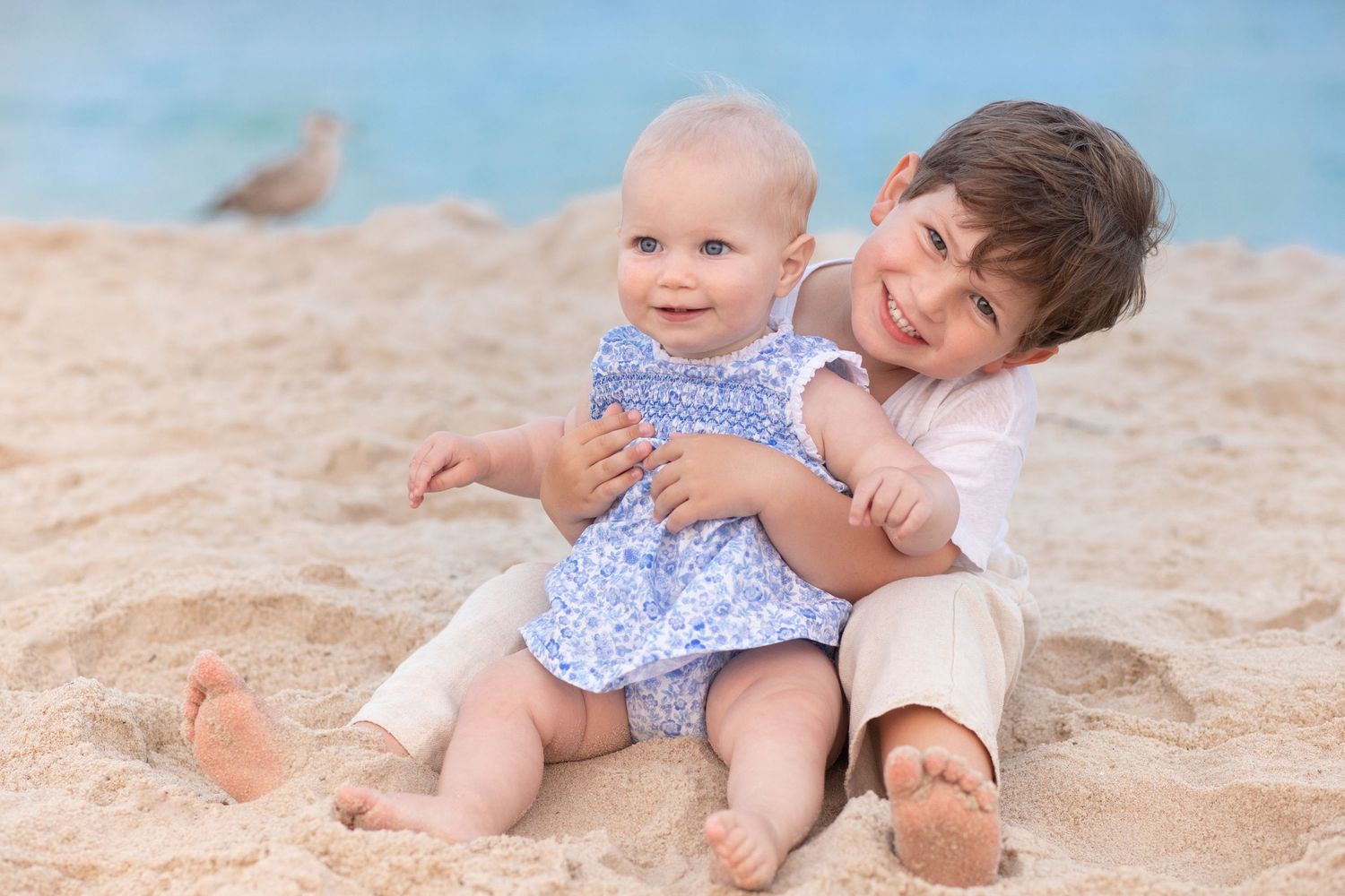 Two young children sitting close together on a sandy beach with blue sky in the background.