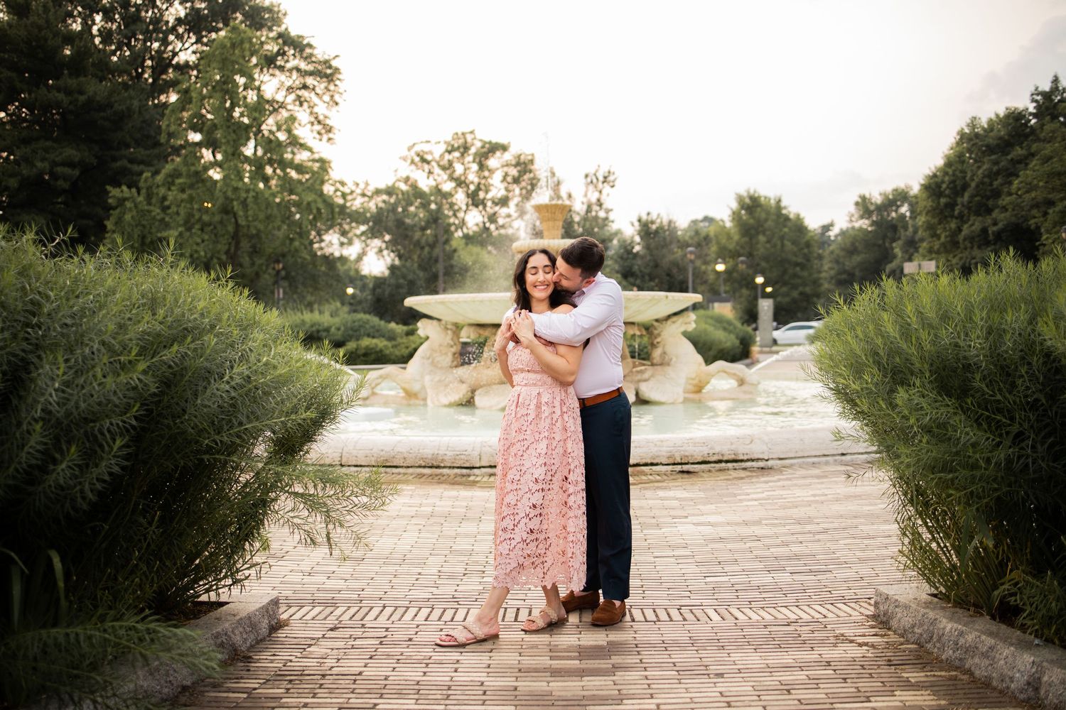 A romantic couple embraces on a garden path lined with lush greenery during golden hour sunset.