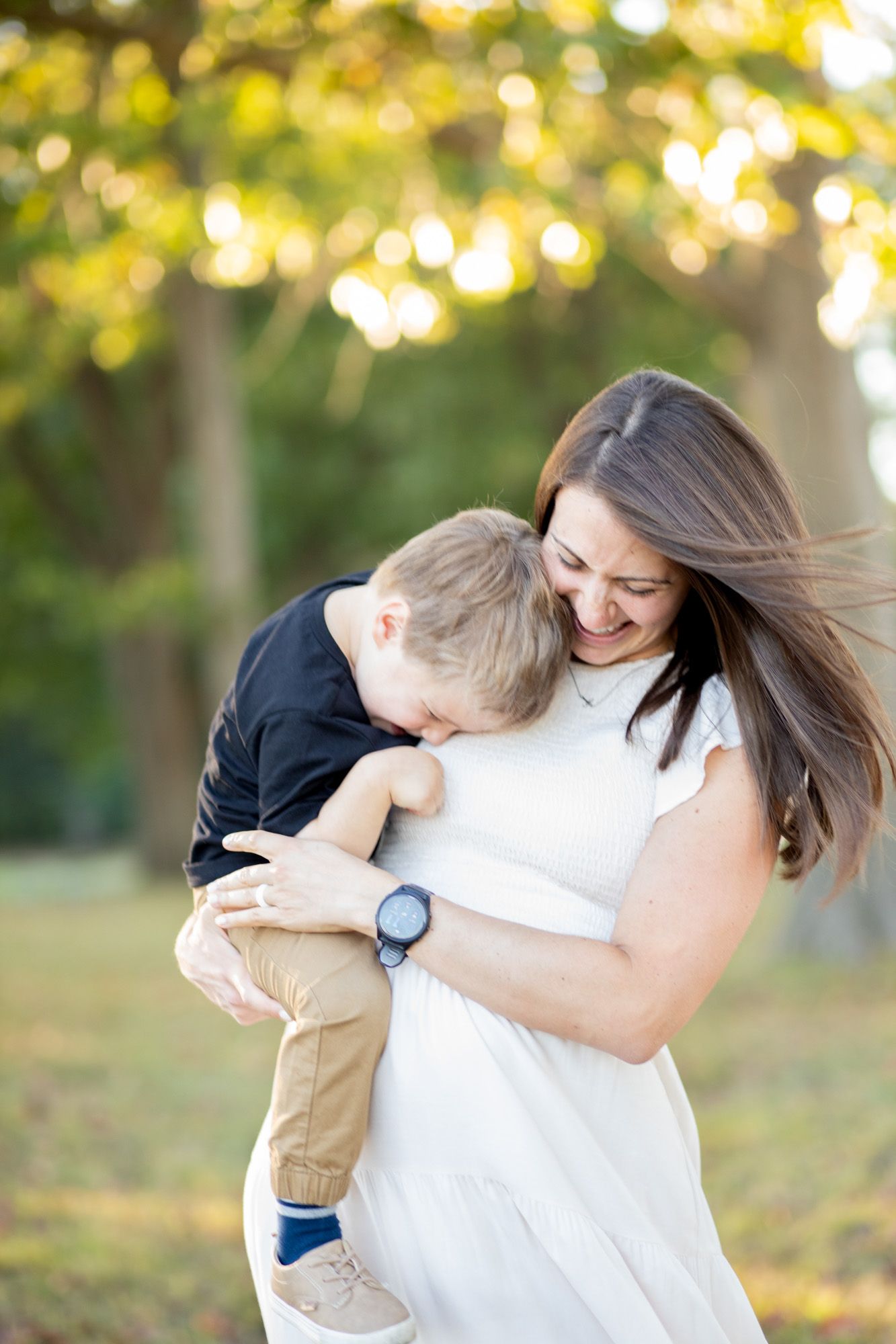 A smiling pregnant woman in a white dress holds a young toddler boy outdoors in a sunlit park.