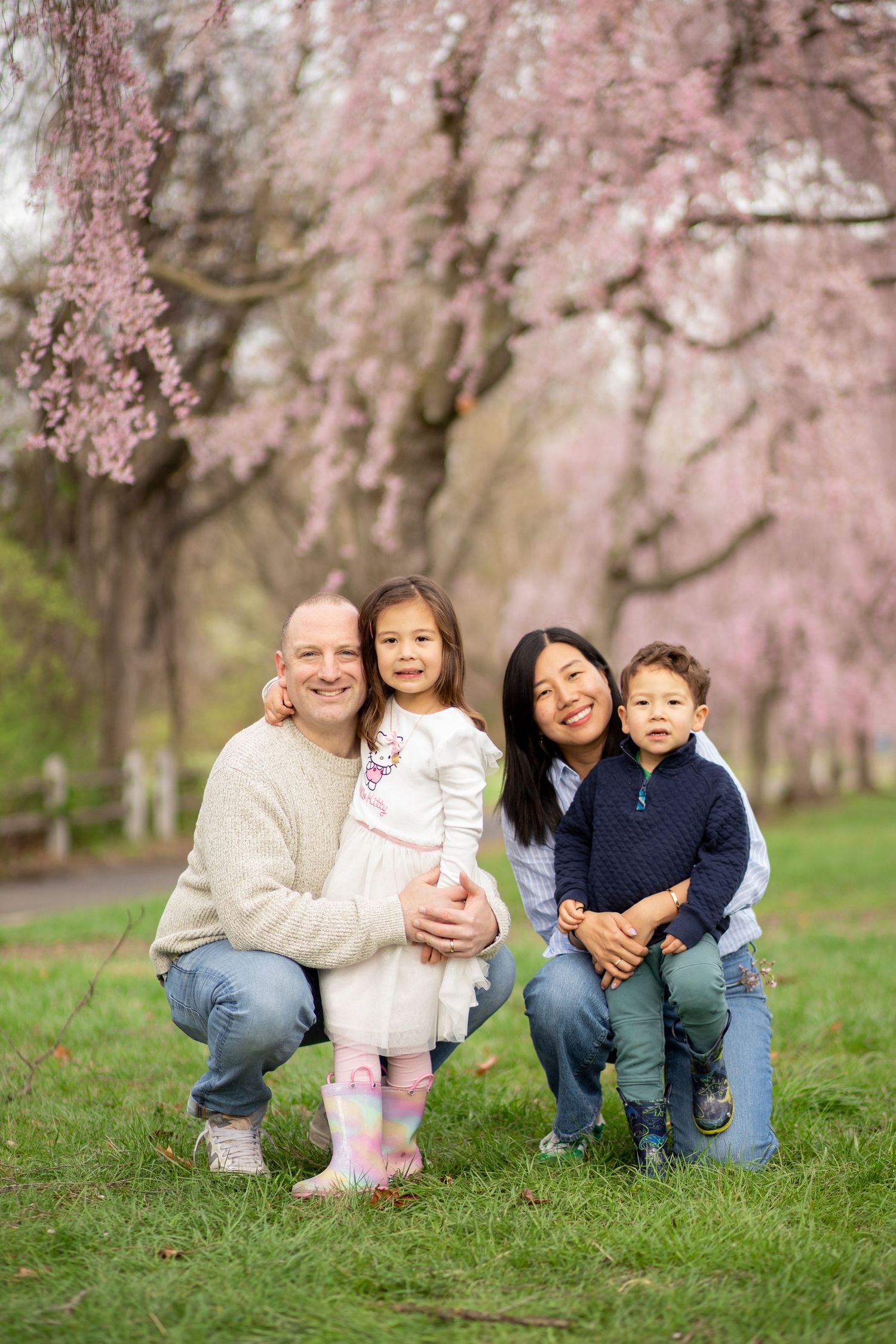 A family poses together under blooming pink cherry blossom trees in a spring garden setting.