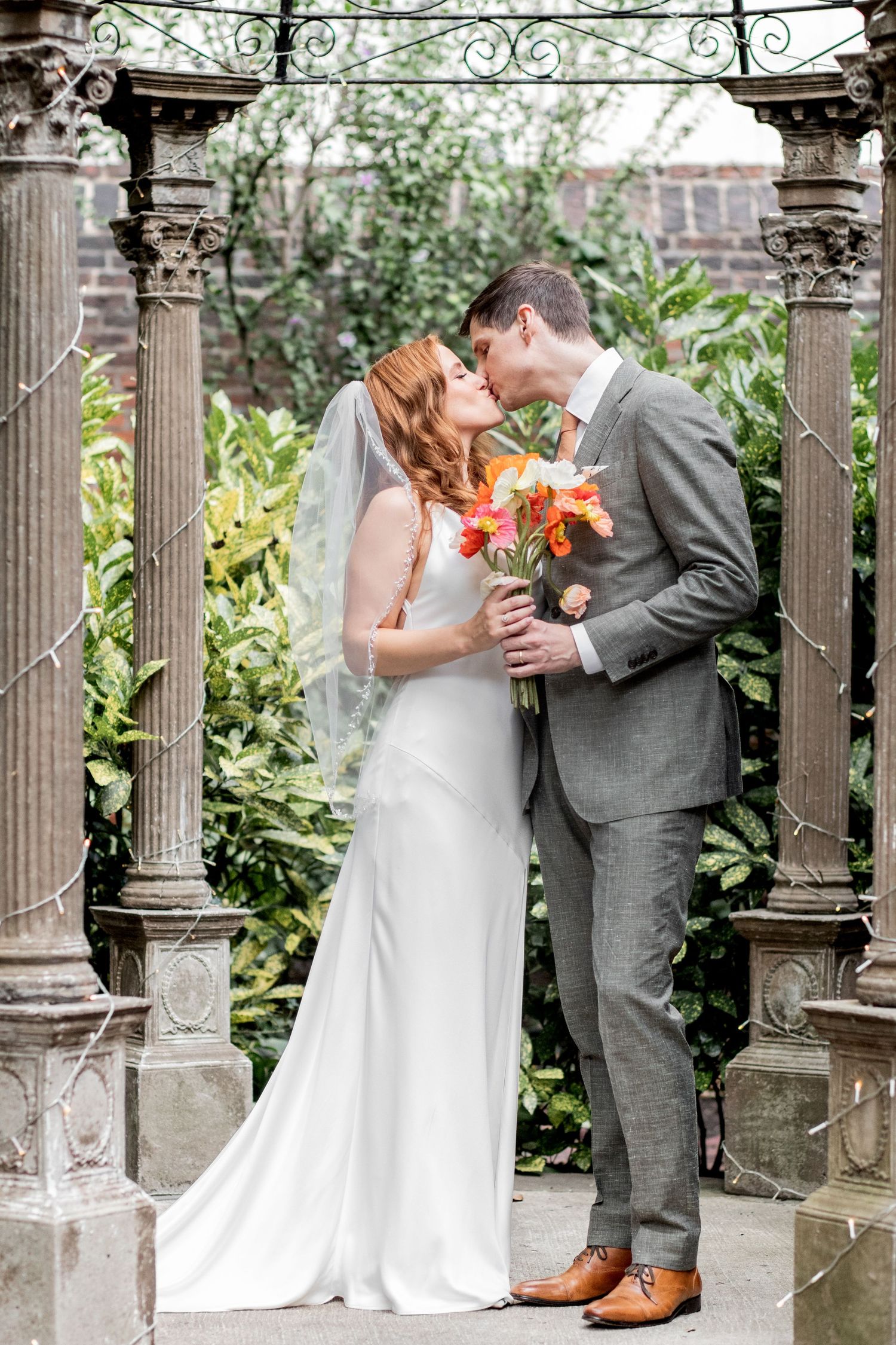 Wedding couple embraces under decorative stone archway with orange bouquet in garden setting.