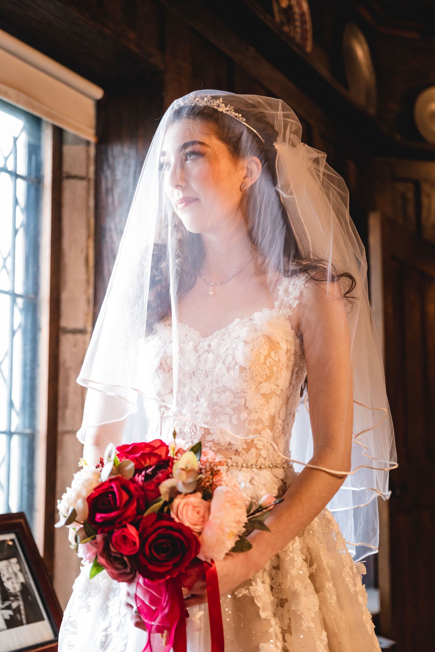 A bride in a white lace wedding dress holds a vibrant bouquet of red and pink roses near a window.