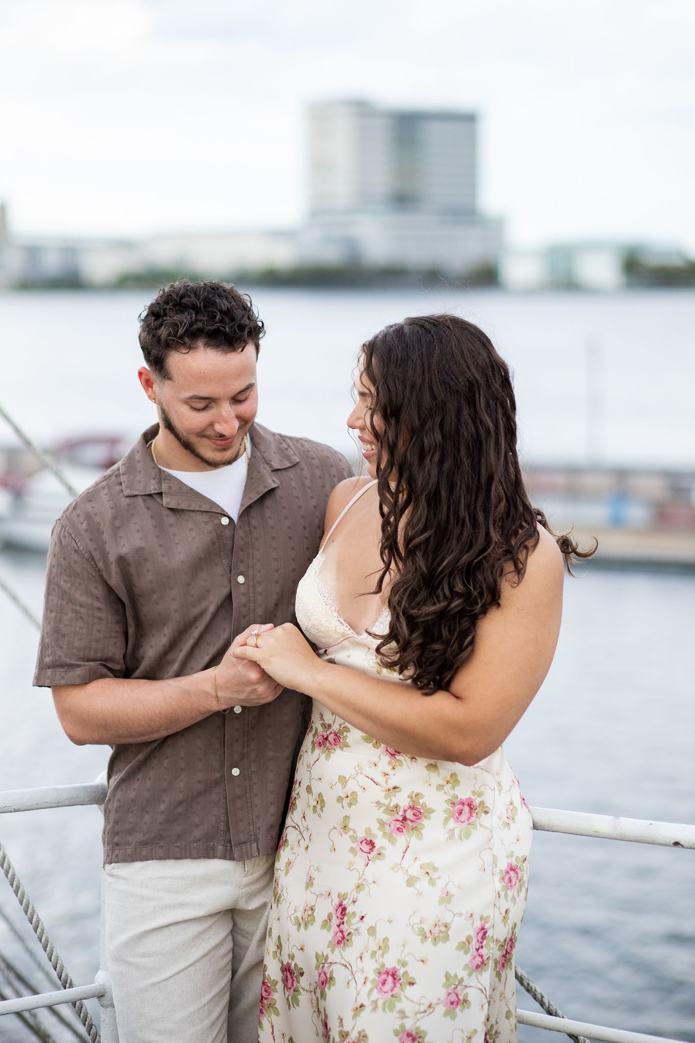 A couple shares a romantic moment on a boat deck overlooking the harbor with buildings in the background.