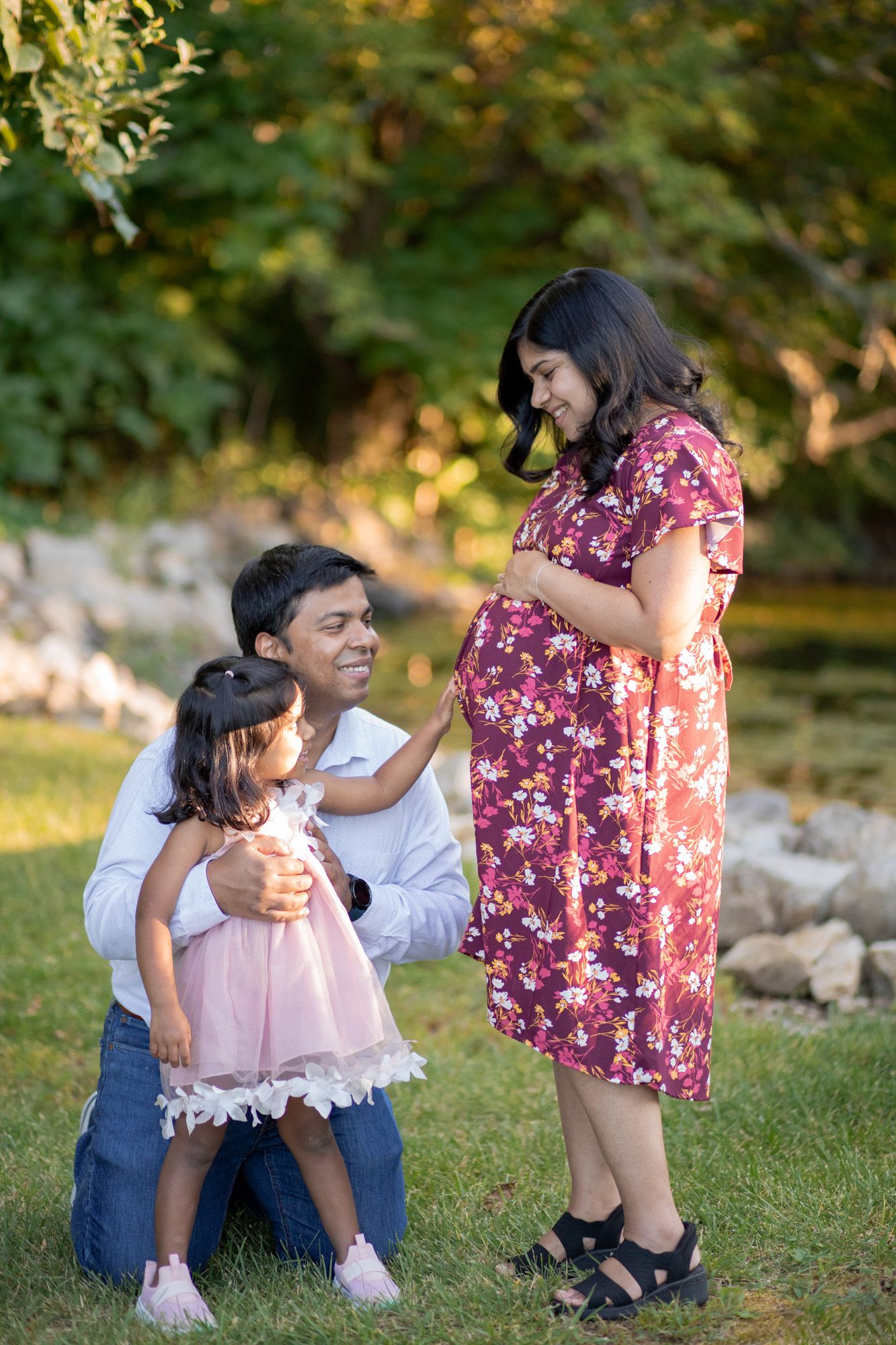 A pregnant woman in a floral dress poses with her husband and toddler daughter in a sunny outdoor park setting.