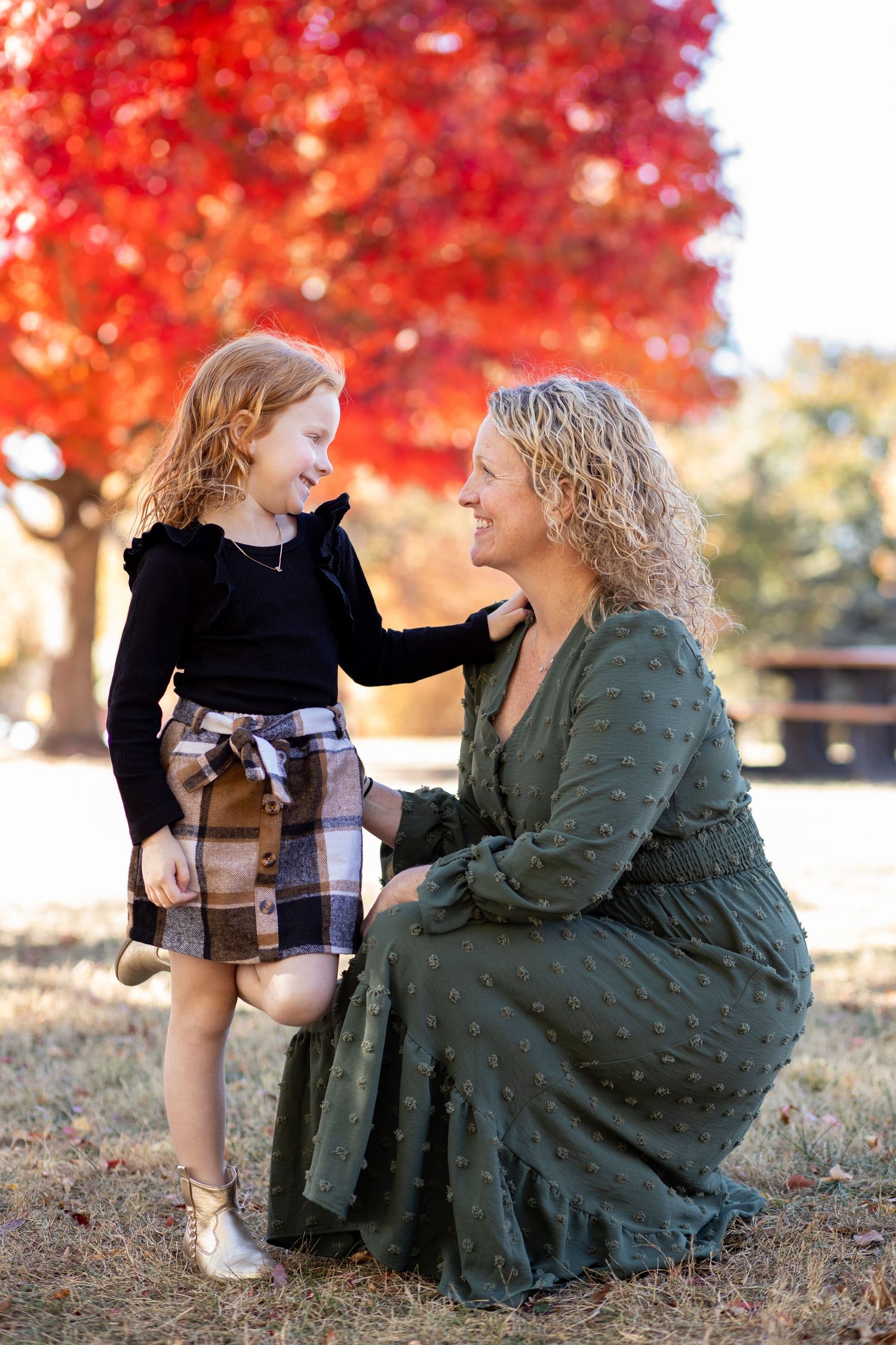 Autumn portrait session featuring tender interaction between figures against vibrant red maple trees and sunlit backdrop.