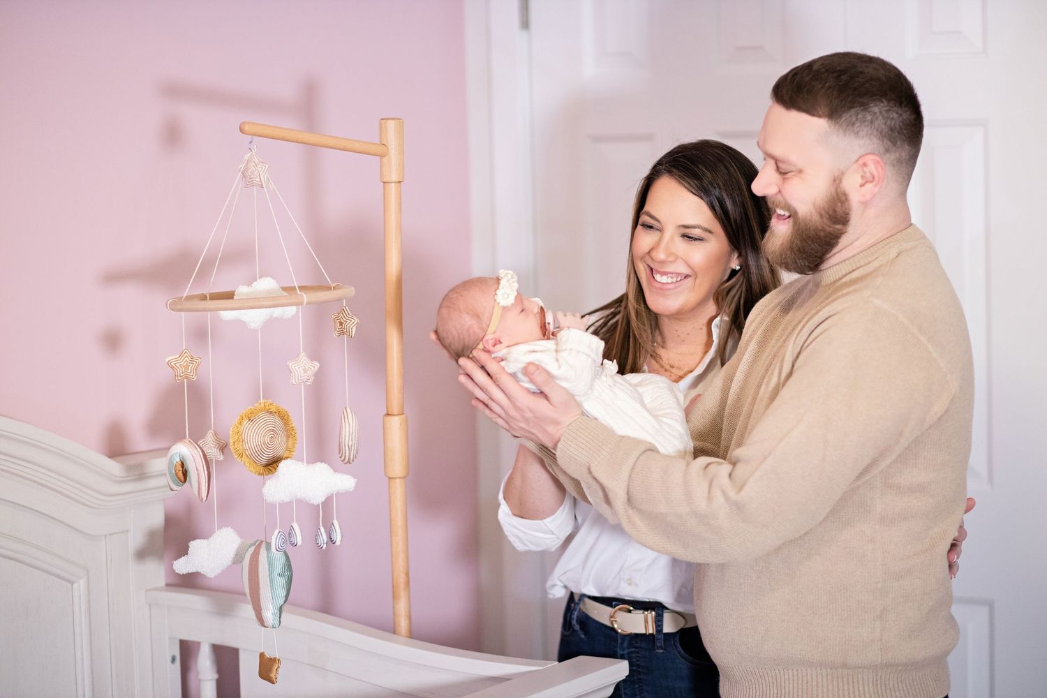 Parents standing near baby mobile in nursery with pink walls as they lovingly hold their newborn.