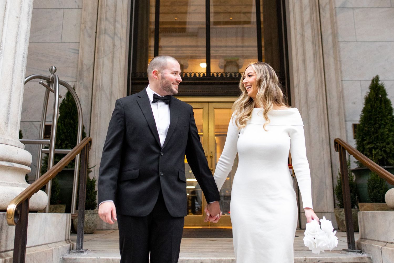 An elegant wedding couple poses in front of a grand stone building entrance with white columns.
