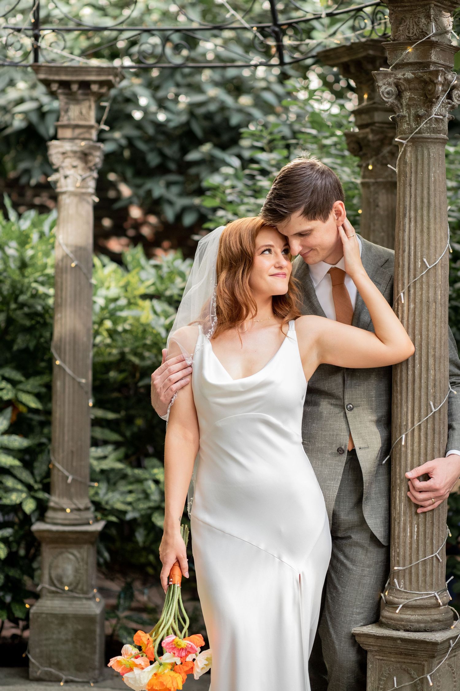 A romantic couple embraces among ornate garden columns, with the person in white holding a vibrant bouquet of flowers.