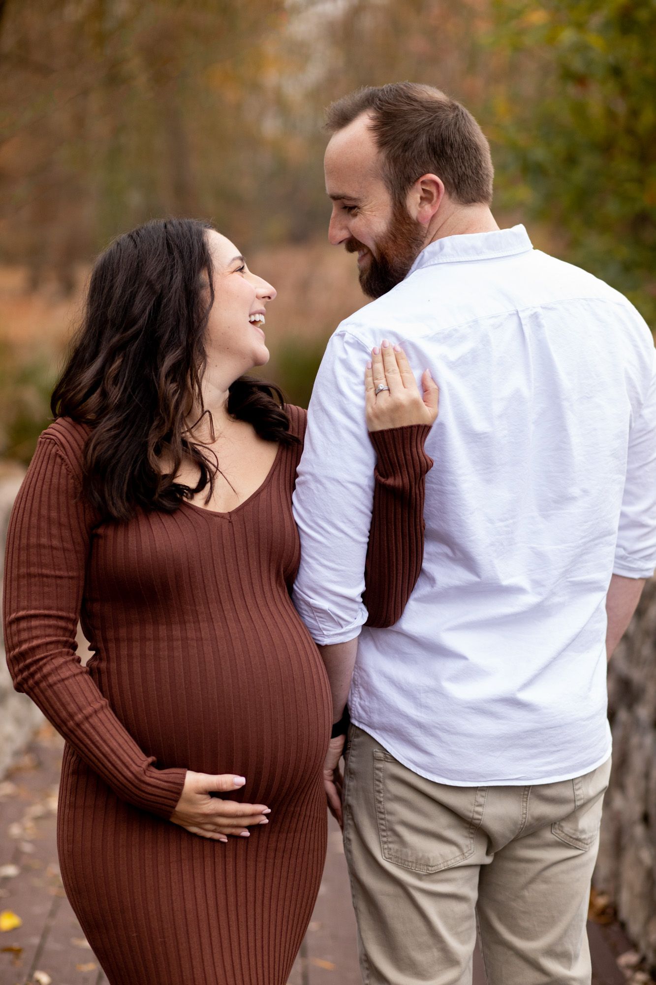 Smiling pregnant woman in brown dress with bearded partner outdoors in autumn park setting.
